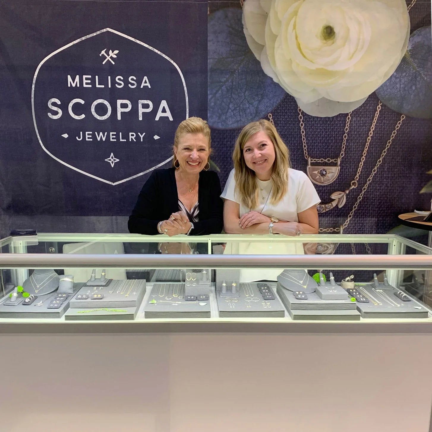 Two women smiling behind glass display case with elegant Melissa Scoppa Jewelry, warm indoor lighting