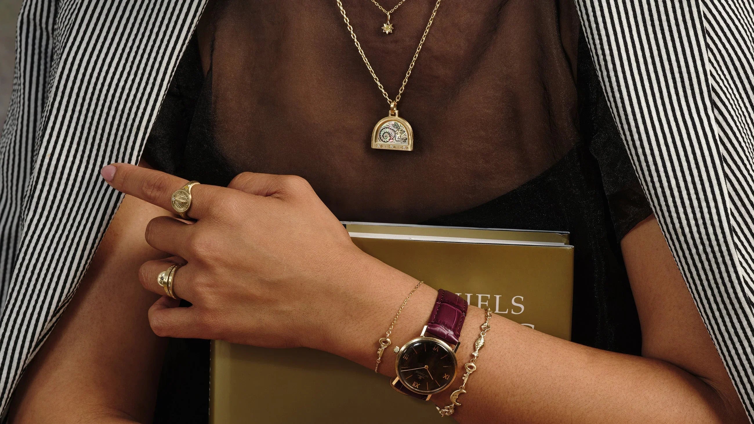 Woman wearing delicate gold sundial ring, engraved gold pendant, and classic watch with purple leather strap in warm natural light