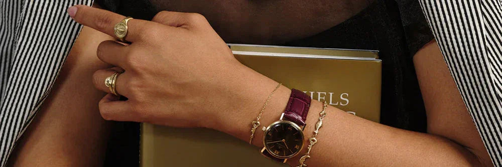 Woman's hand wearing elegant gold rings and watch with burgundy strap, holding a gold book, soft warm lighting