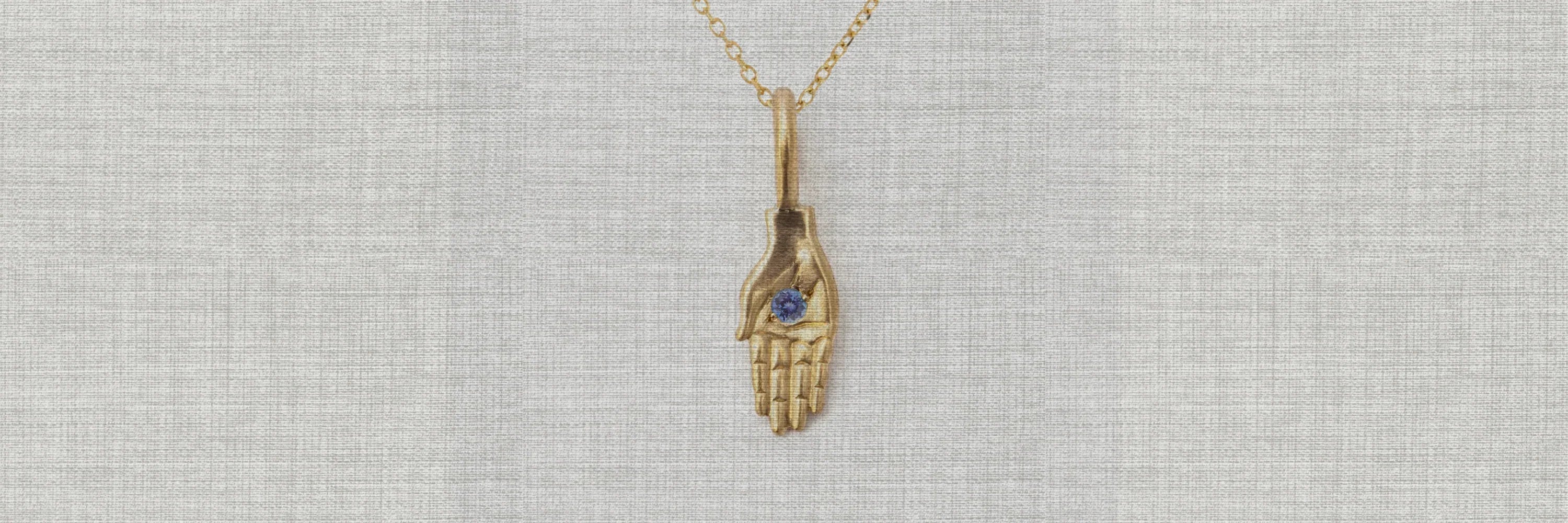 Hand-fabricated gold hand pendant with blue birthstone on textured neutral linen background