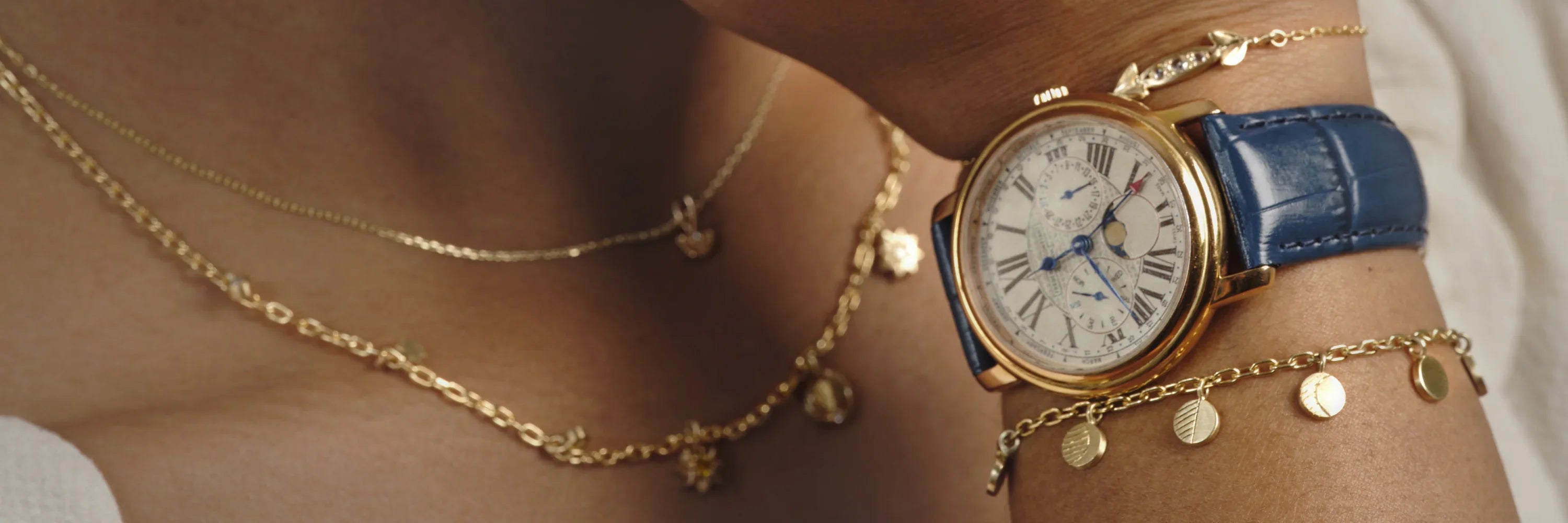 Close-up of wrist with gold charm bracelet and vintage gold watch with blue leather strap on warm skin tone
