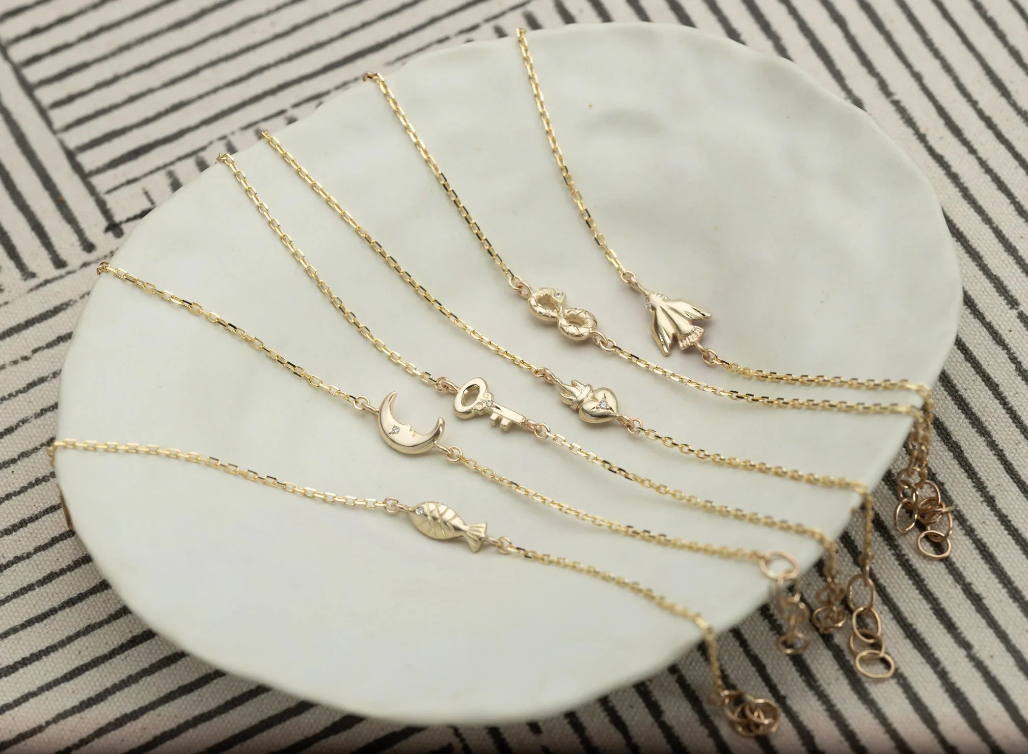 Delicate 14k gold charm bracelets with moon, key, heart, infinity, fish, and flower motifs on white ceramic plate