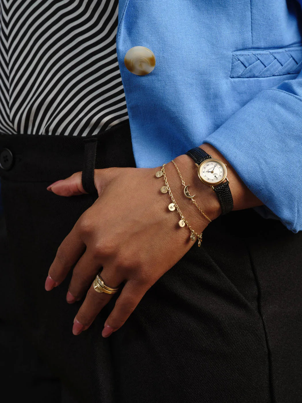 Close-up of wrist with vintage gold watch and delicate gold moon phase and charm bracelets, styled with blue blazer