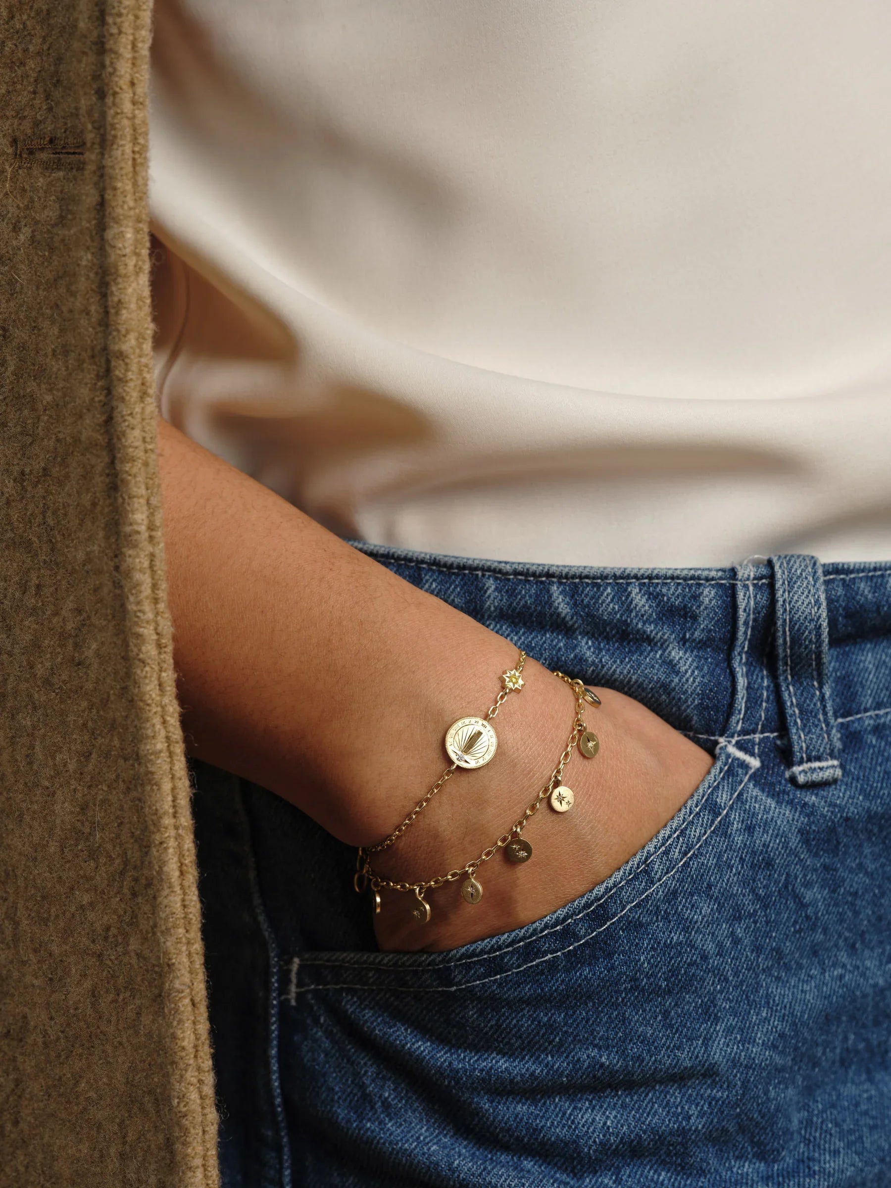 Wrist with two delicate gold bracelets: a sundial charm and a chain with small celestial charms, natural light