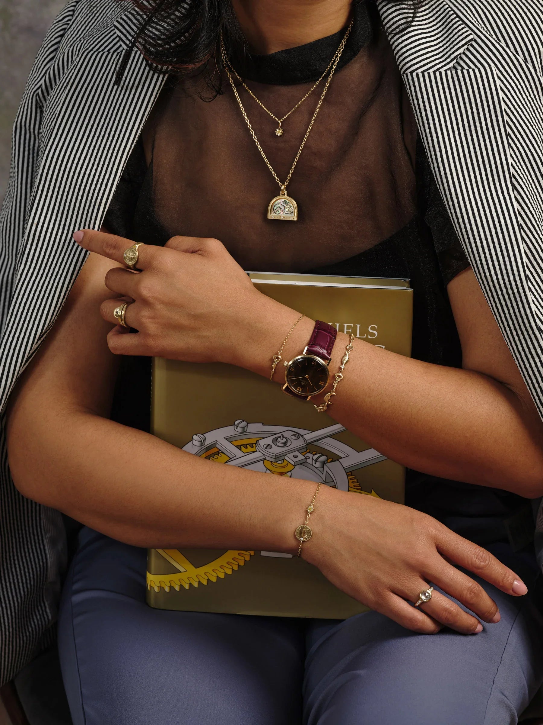 Woman wearing layered gold necklaces, rings, and moon phase bracelets holding a horology book in warm natural light