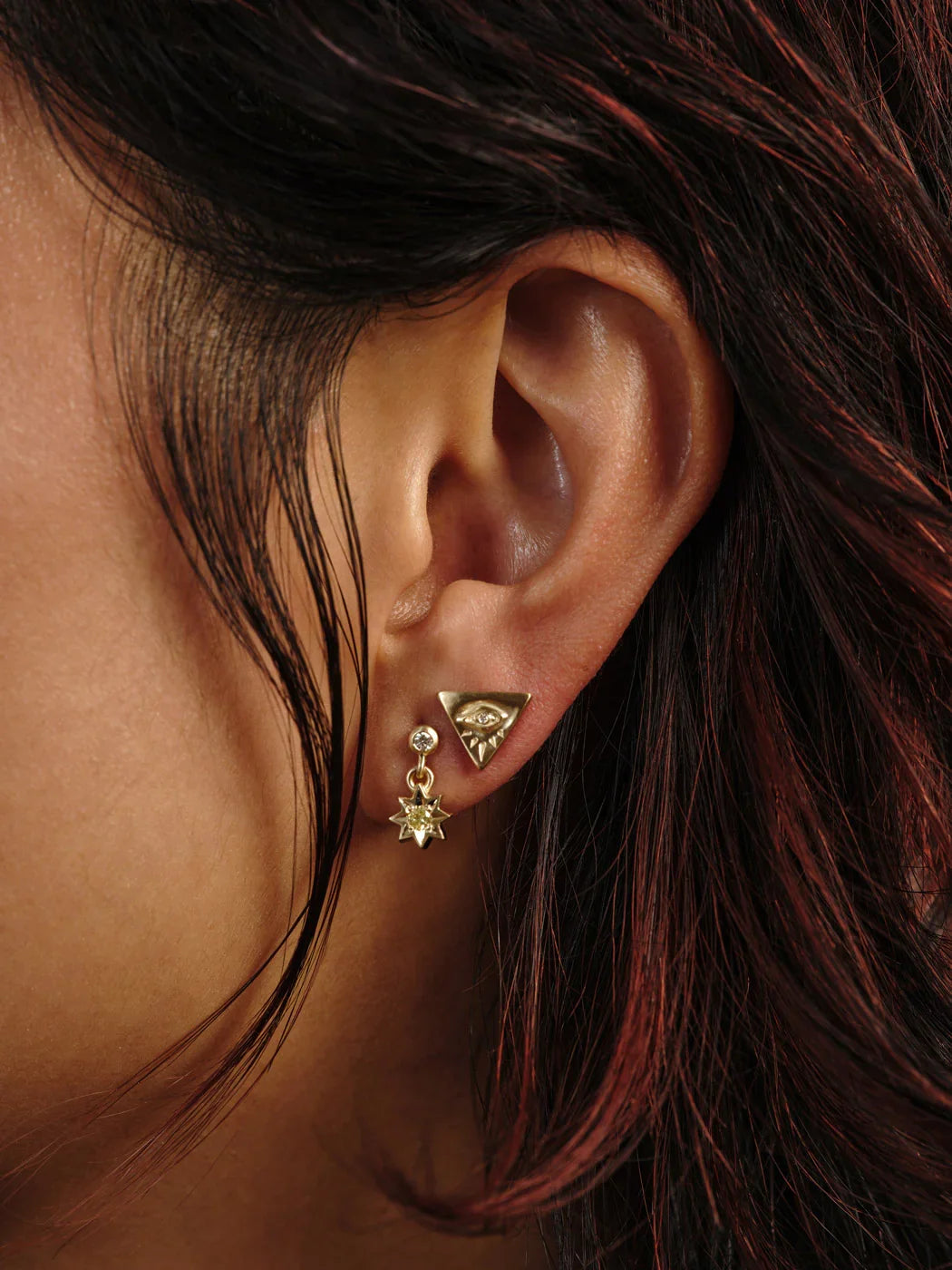 Close-up of ear wearing hand-fabricated gold eye and star earrings with warm natural light