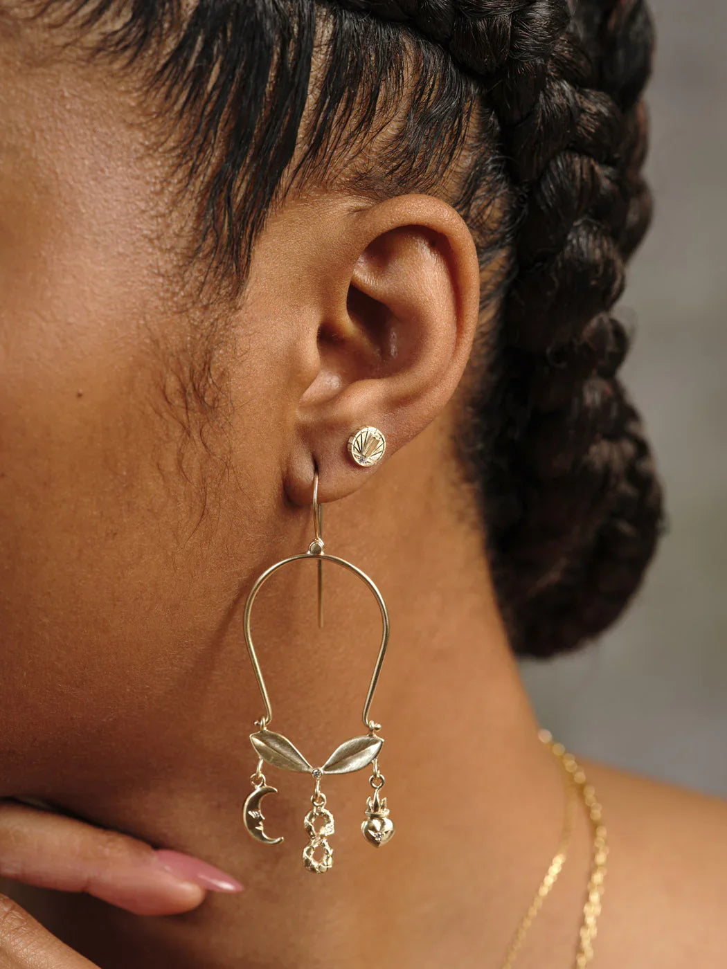 Close-up of woman's ear wearing elegant gold earrings with moon, floral, and heart charms against neutral background