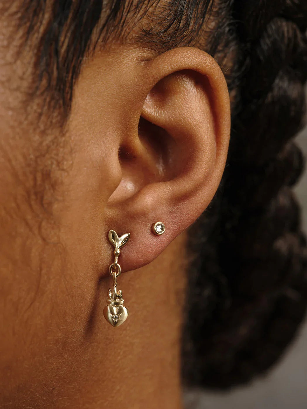 Close-up of ear wearing gold heart and leaf drop earring with small stud, warm natural lighting