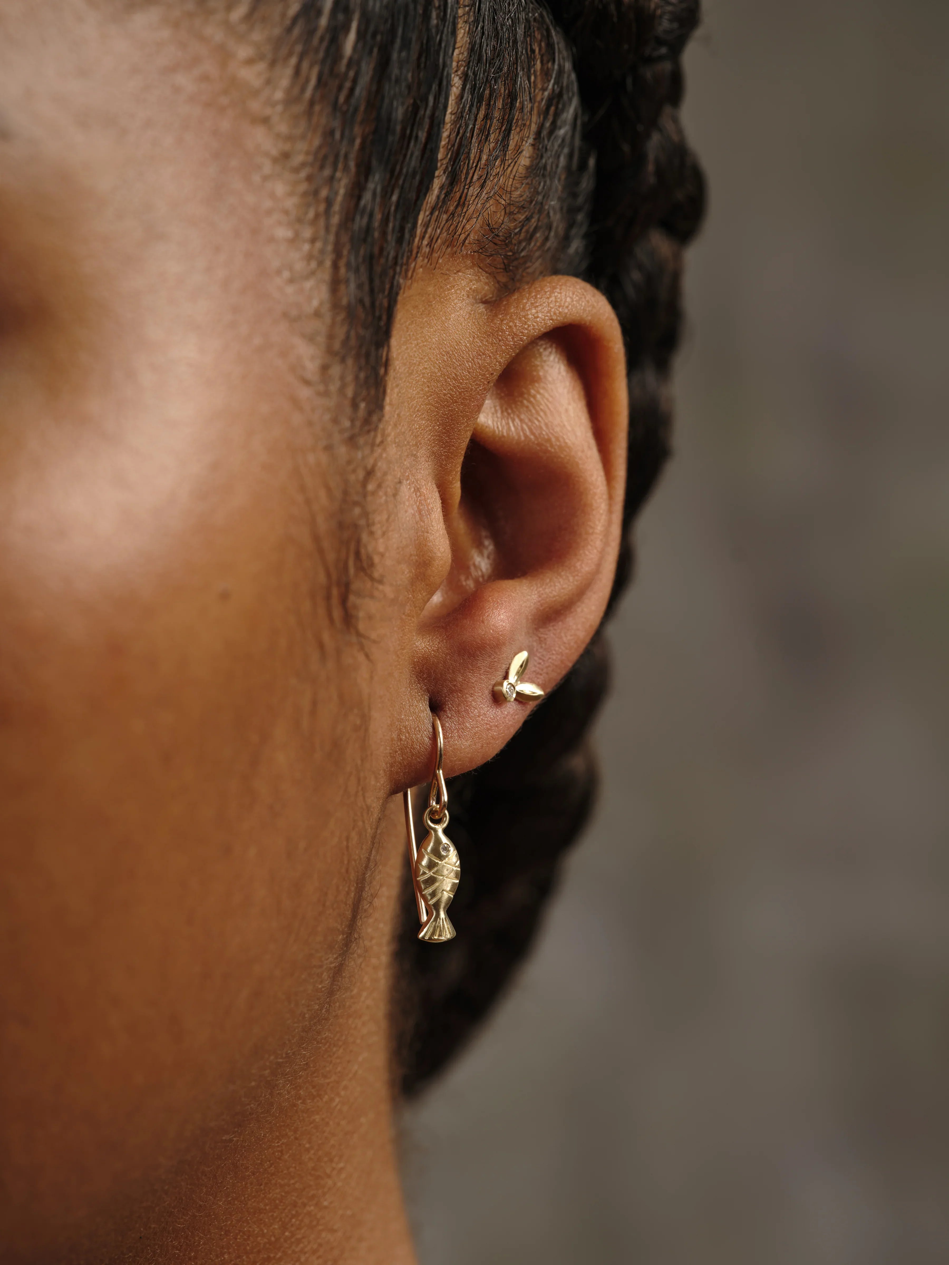 Close-up of ear wearing handcrafted gold fish charm drop earring and small gold triple petal stud against blurred neutral background