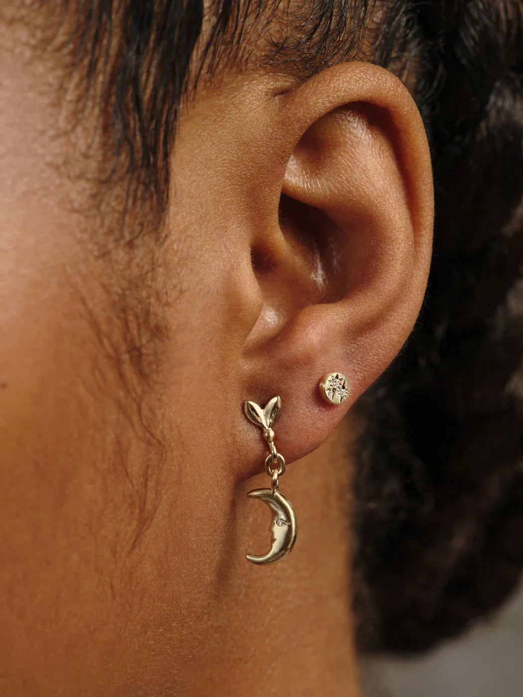 Close-up of ear wearing handcrafted gold moon phase dangle earring and small star-studded gold stud, warm natural light