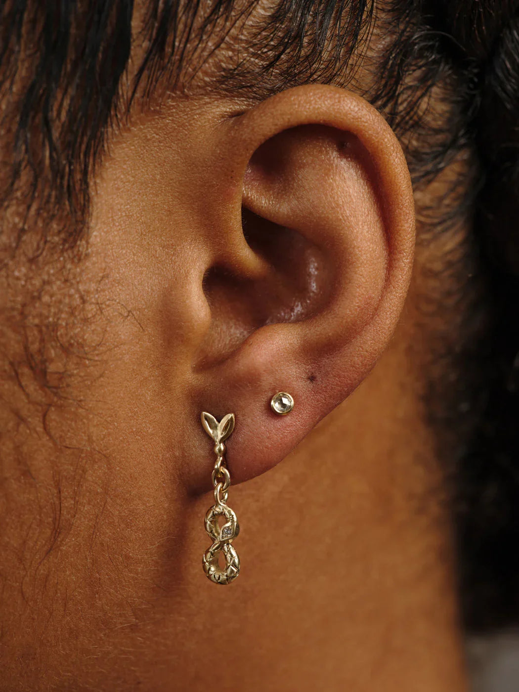 Close-up of ear with delicate hand-fabricated Fairmined gold dangle earring and small gold stud, warm natural light