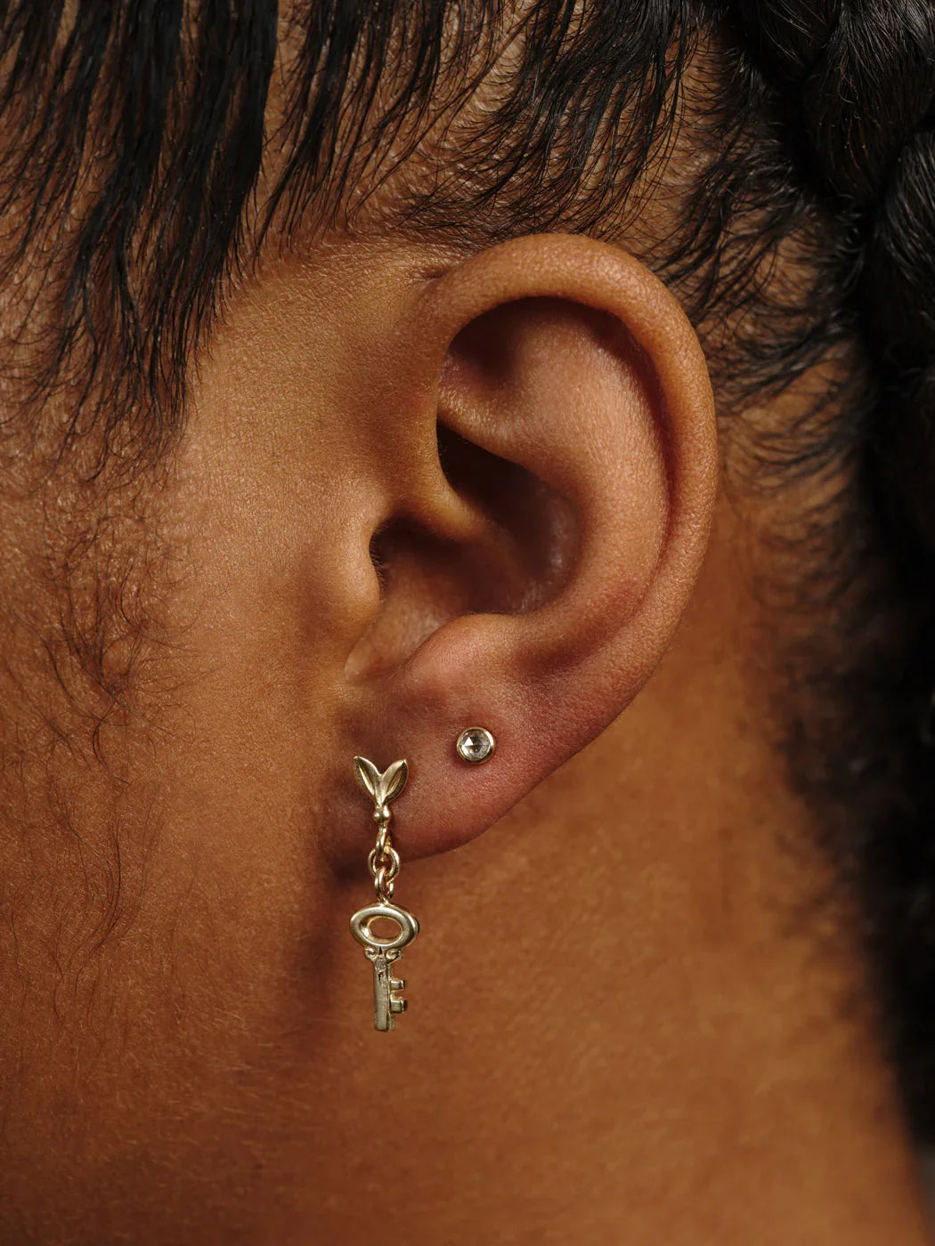 Close-up of ear wearing delicate gold key drop earring and small gold stud in warm natural light