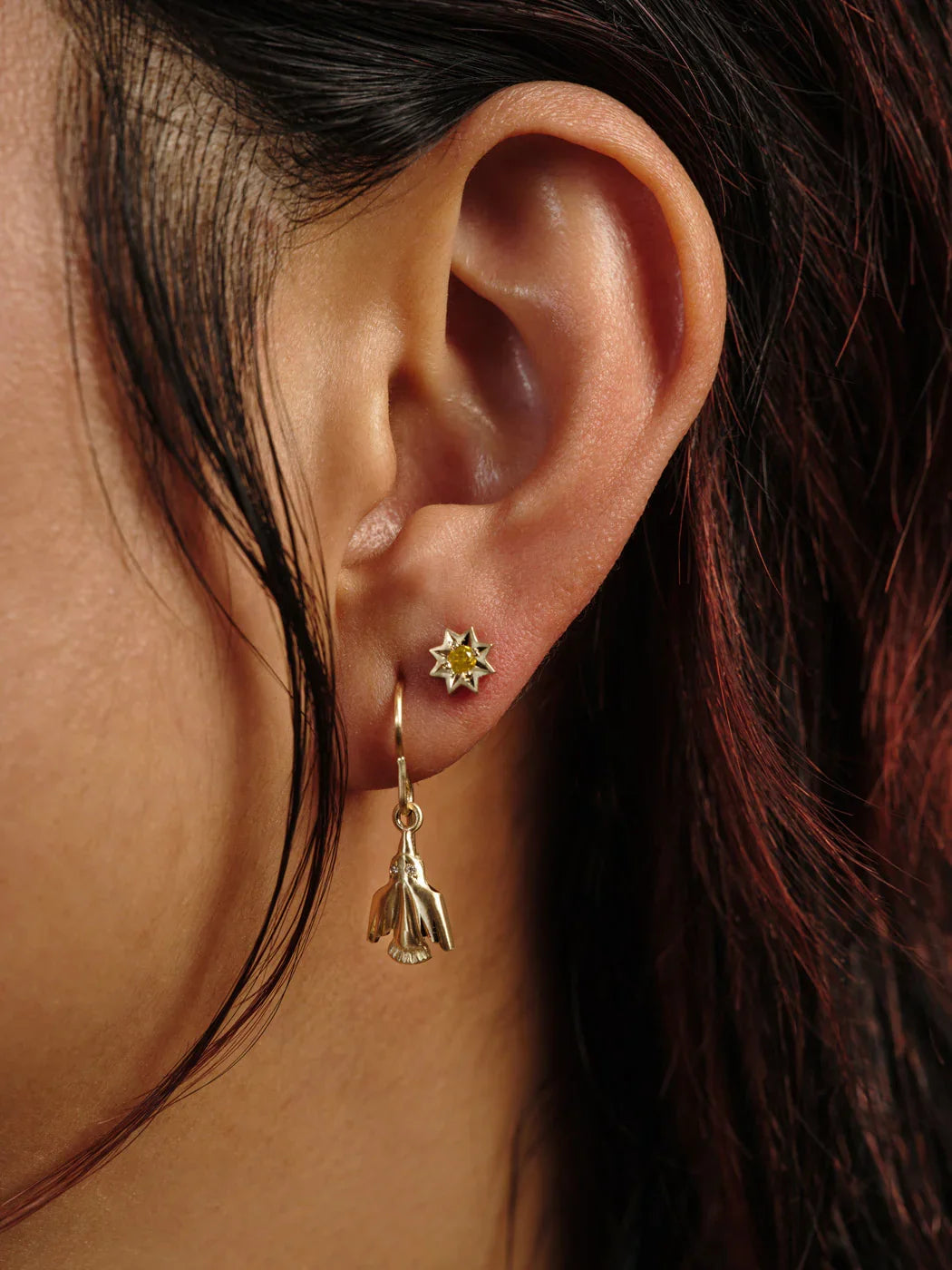 Close-up of ear with hand-fabricated gold sunburst stud and bird-shaped gold drop earring, warm natural light