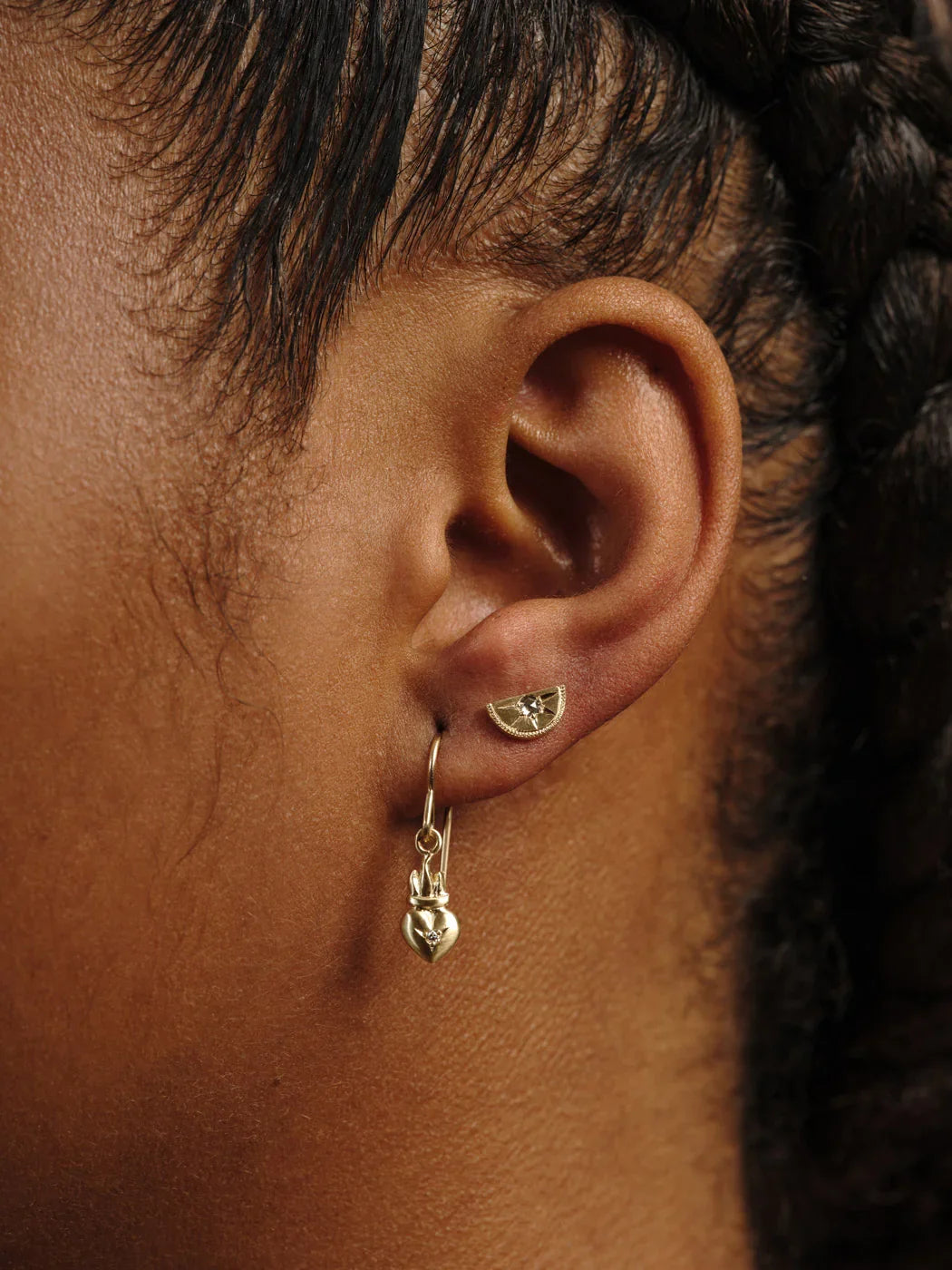 Close-up of ear wearing handcrafted gold half-moon stud and heart-shaped dangle earring in warm natural light