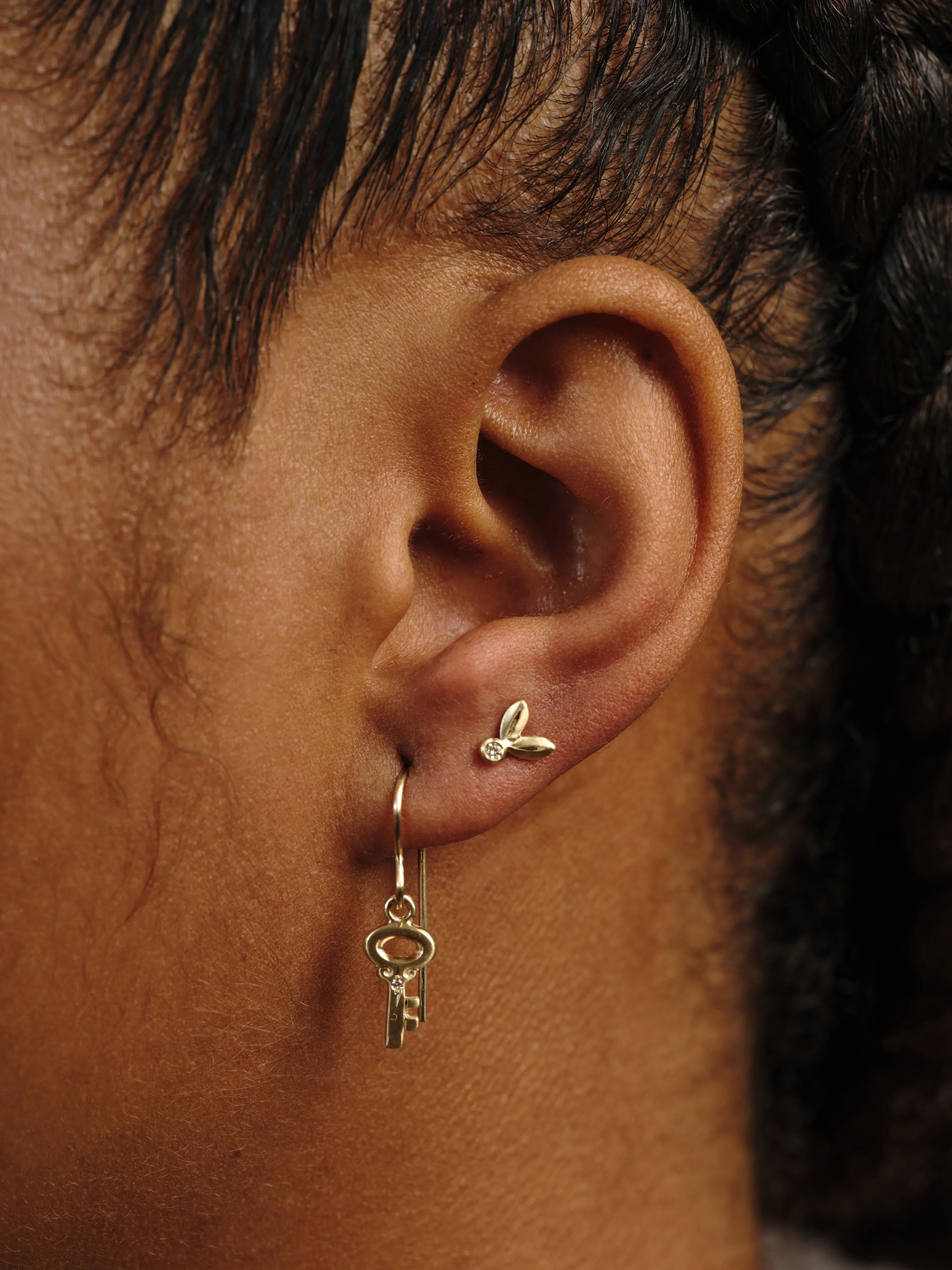 Close-up of ear wearing delicate handcrafted gold earrings, including key charm dangle and small stud with diamond, warm natural light