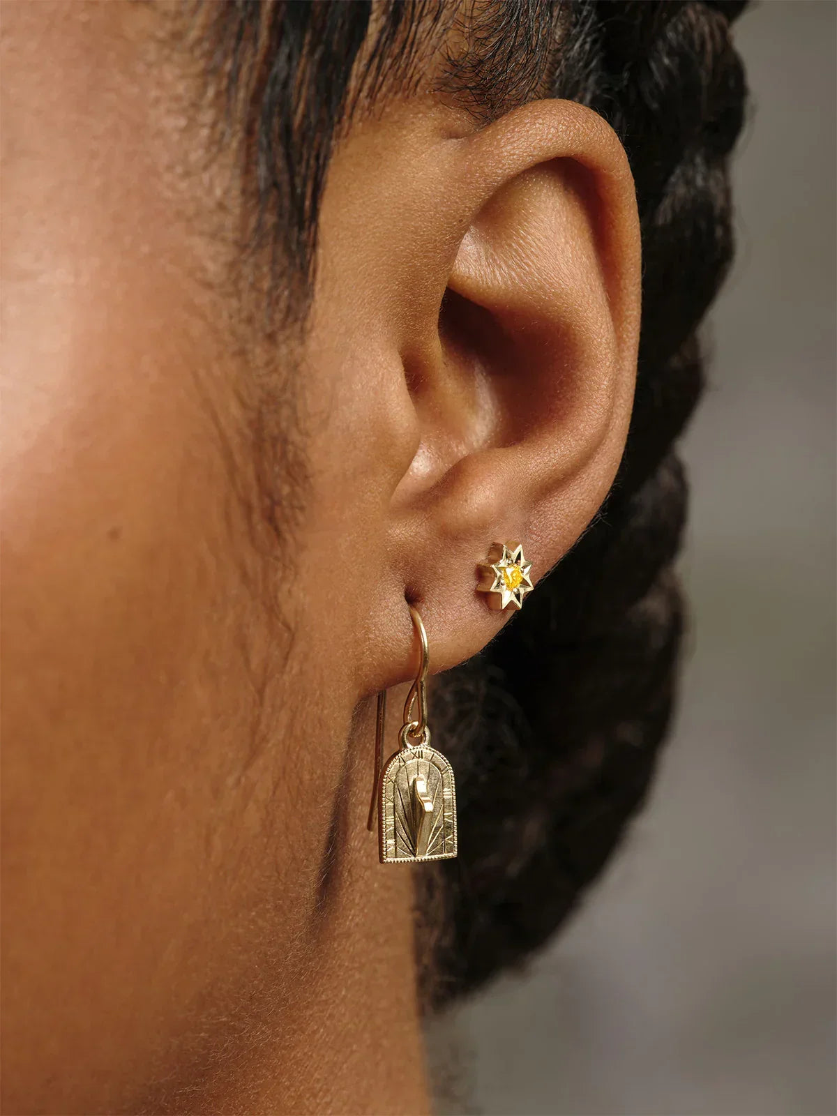 Close-up of ear wearing handcrafted Fairmined gold sundial drop earring and star-shaped stud, warm natural light