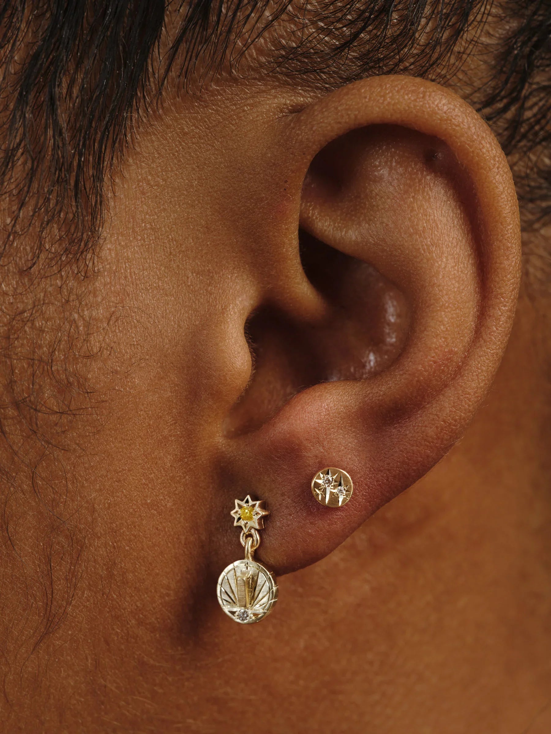 Close-up of ear wearing hand-engraved Fairmined gold sundial and star stud earrings in warm natural light
