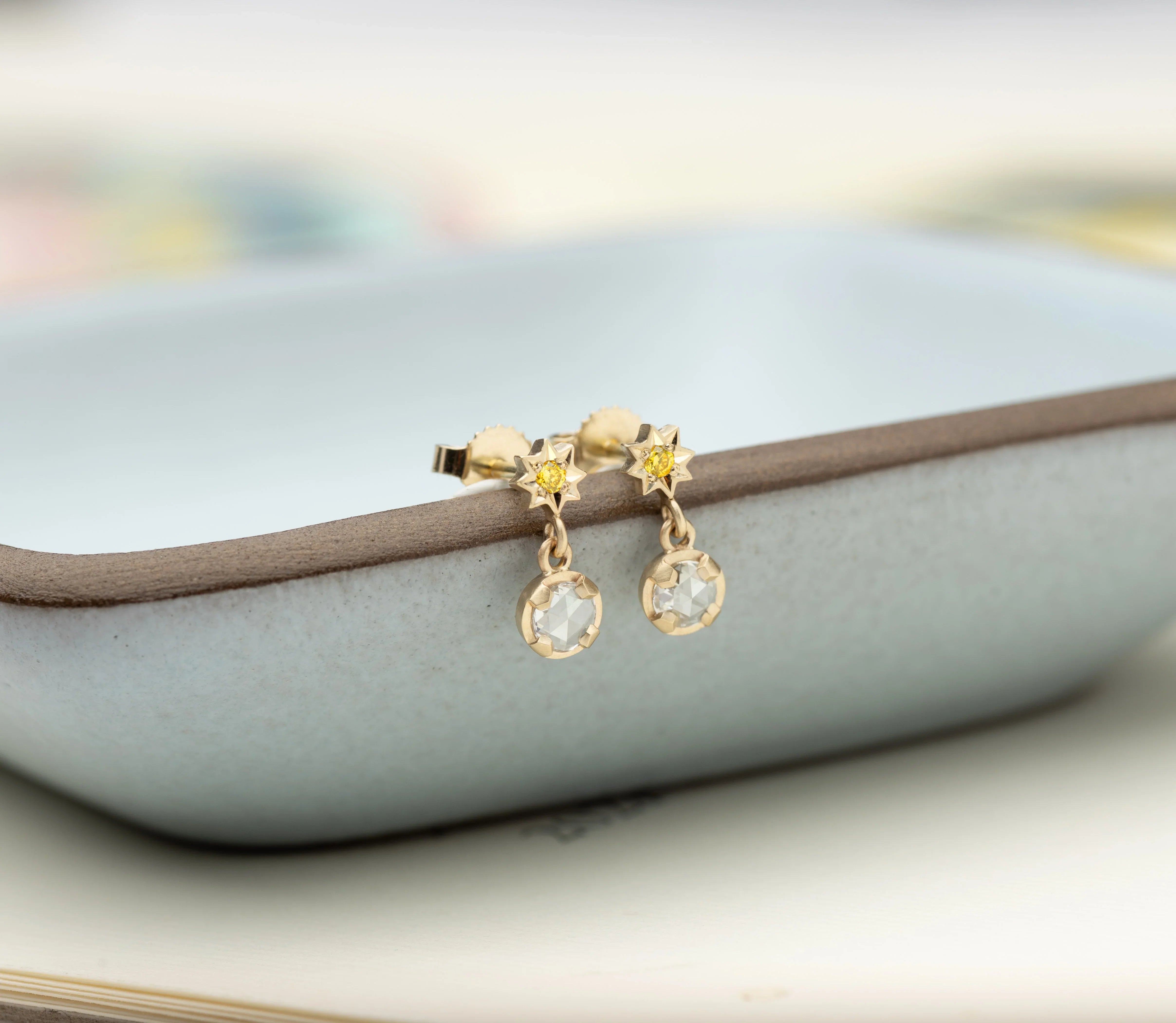 Delicate Fairmined gold star stud earrings with yellow gemstone centers and small hanging clear stones on pale ceramic dish