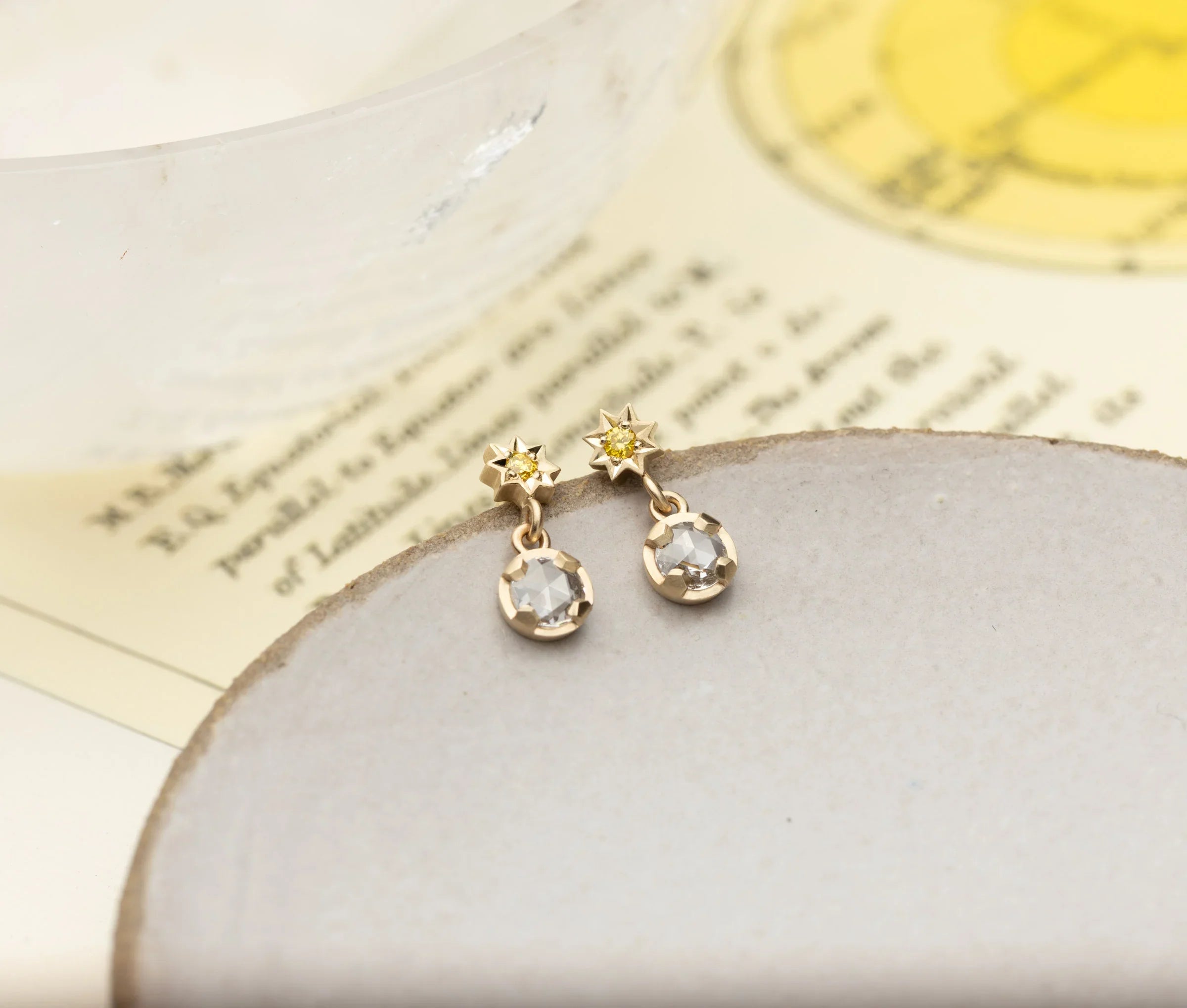 Pair of delicate gold star-shaped earrings with yellow center stones and clear drops on neutral ceramic plate