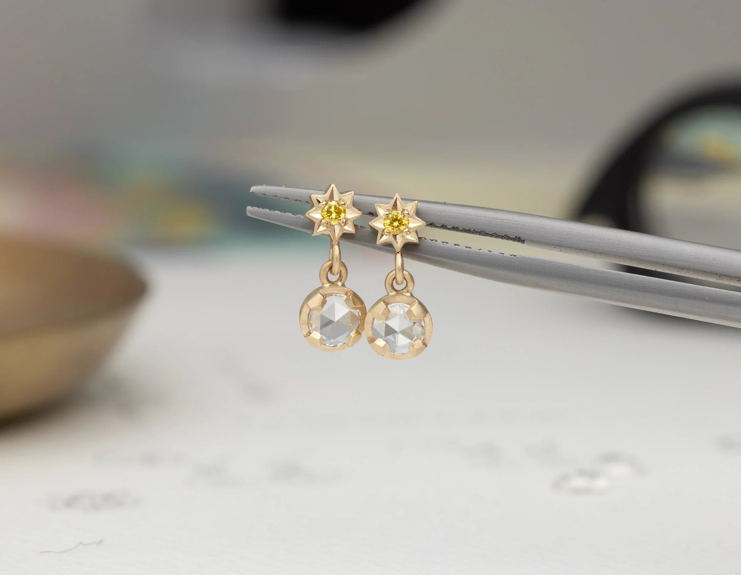 Close-up of gold star stud earrings with yellow center stones and faceted clear drops held by tweezers on soft background