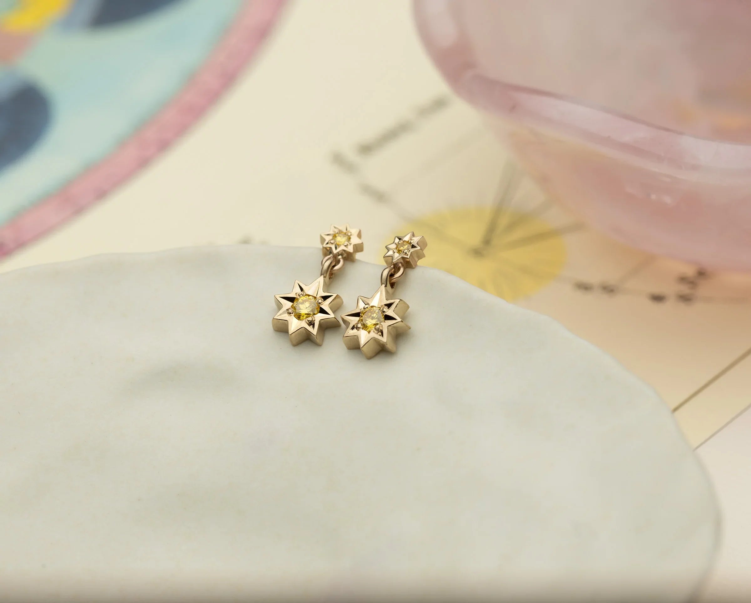 Pair of handcrafted gold star-shaped earrings with yellow gemstones on pale ceramic dish, warm soft light