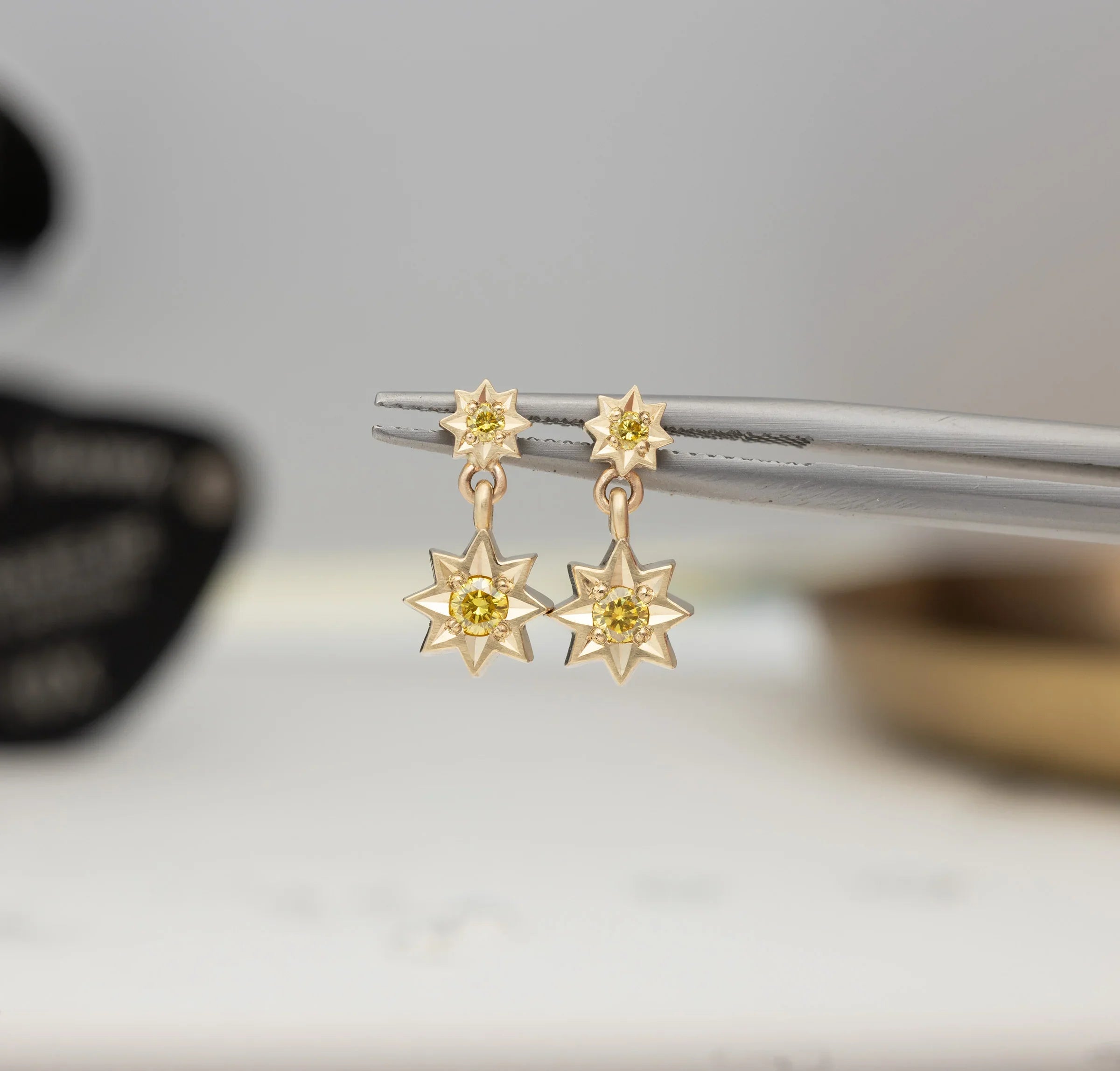 Close-up of handcrafted gold star earrings with yellow gemstones held by tweezers, neutral blurred background