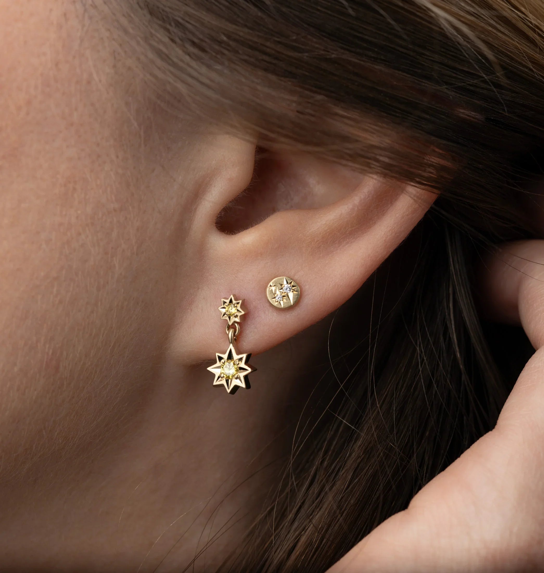 Close-up of ear wearing hand-fabricated gold star-shaped earrings with yellow gemstones, soft natural light