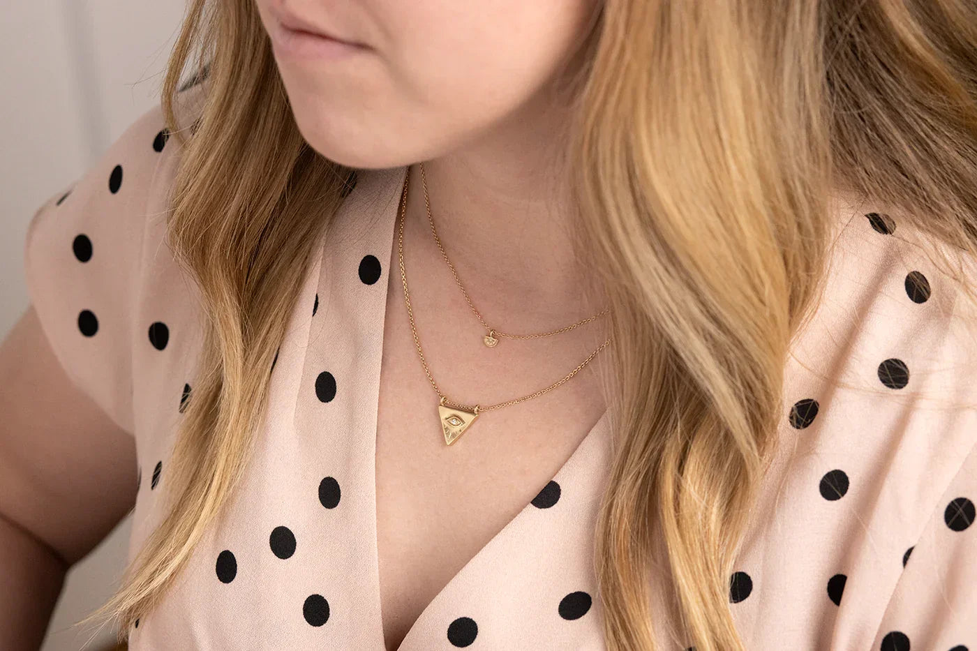 Woman wearing layered gold Evil Eye pendants on delicate chains with soft natural lighting and neutral polka-dot blouse