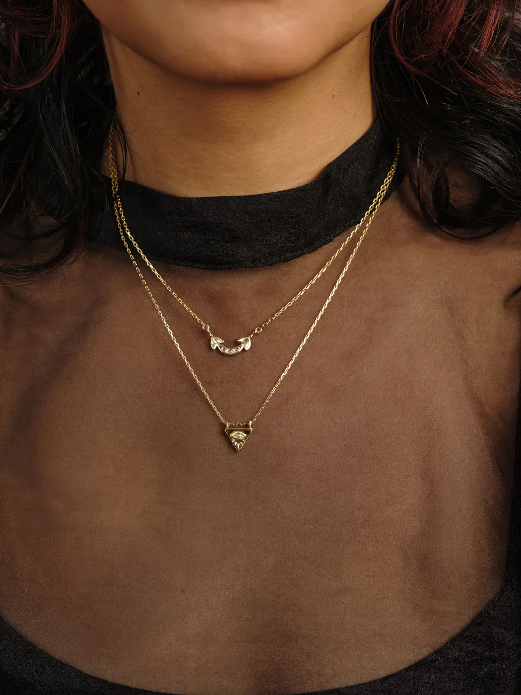 Close-up of layered gold necklaces with crescent and triangle pendants on woman wearing sheer brown top and black collar