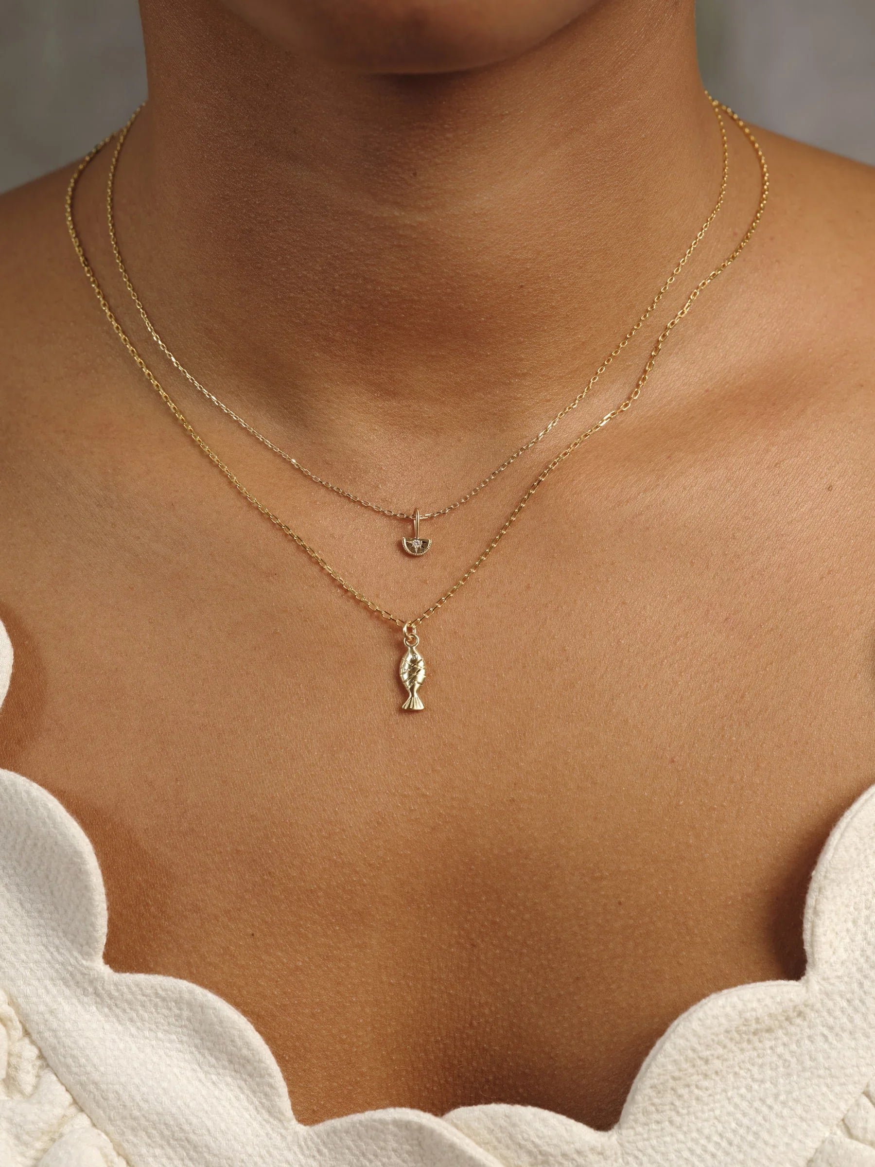 Close-up of two delicate gold necklaces on a woman's neck with warm natural light and neutral scalloped white top