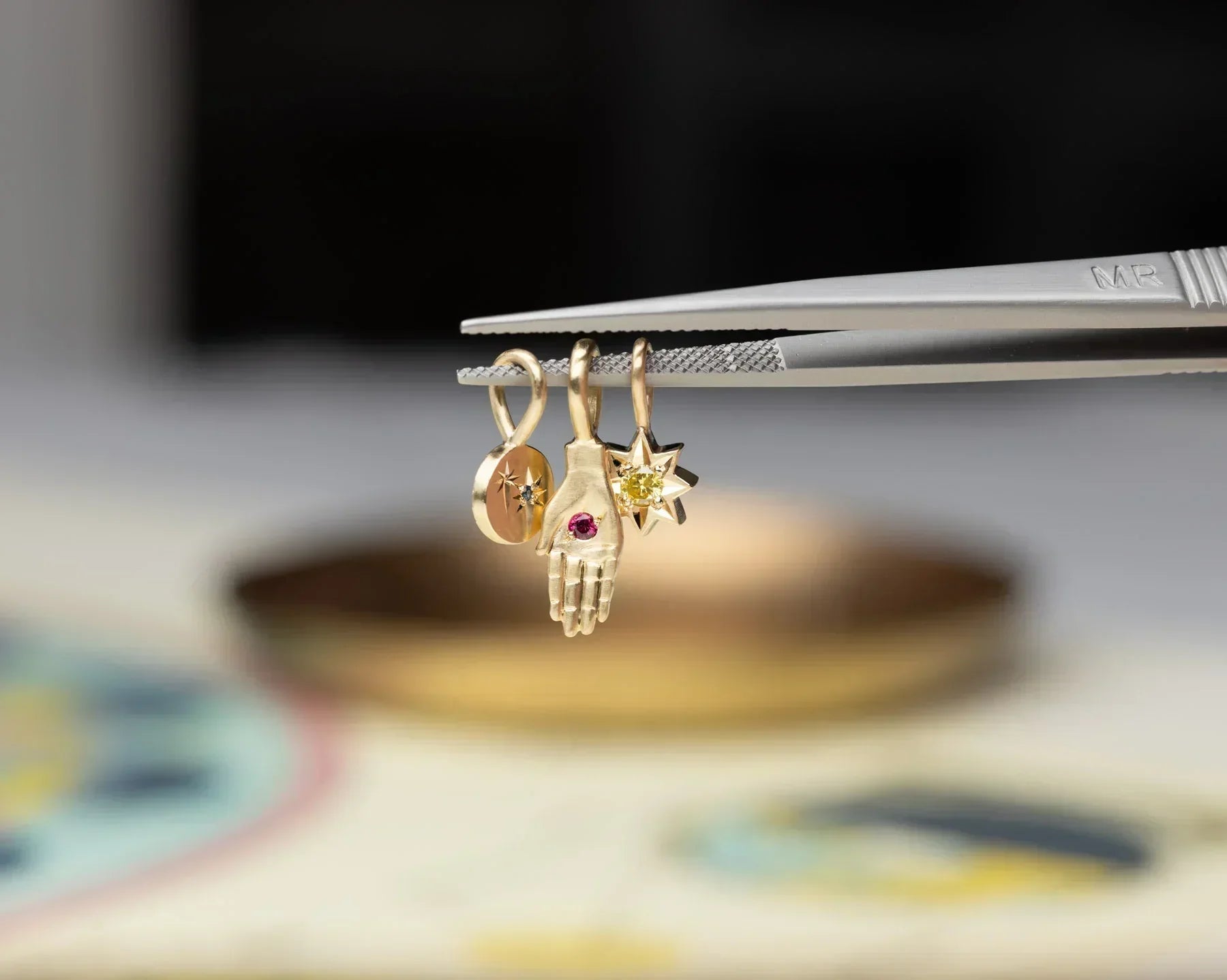 Close-up of three hand-fabricated Fairmined gold charms held by tweezers, featuring a hand with ruby, star with yellow gemstone, and engraved disc, soft warm lighting
