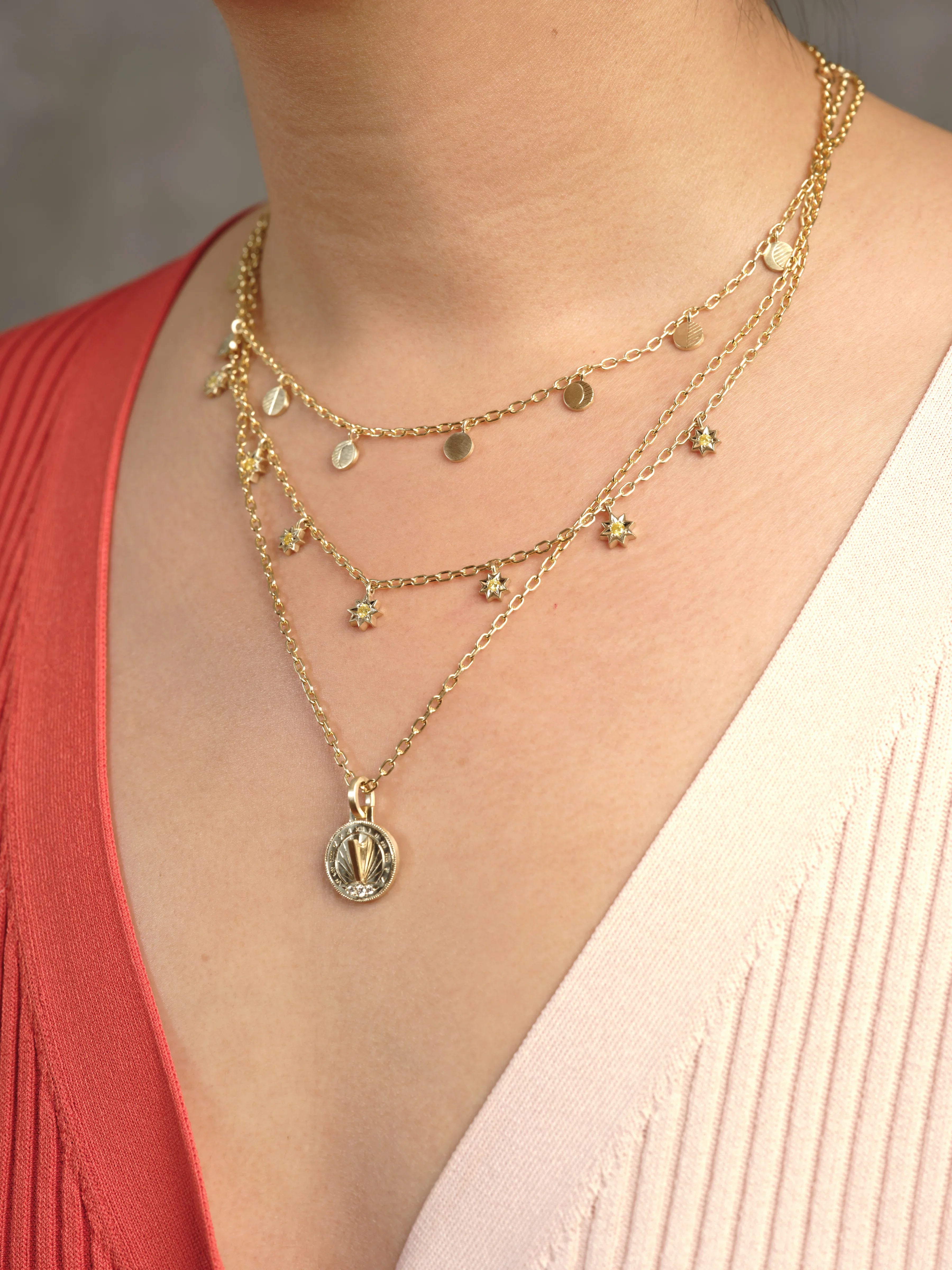 Close-up of layered Fairmined gold necklaces with sun and disc charms on skin, warm natural light