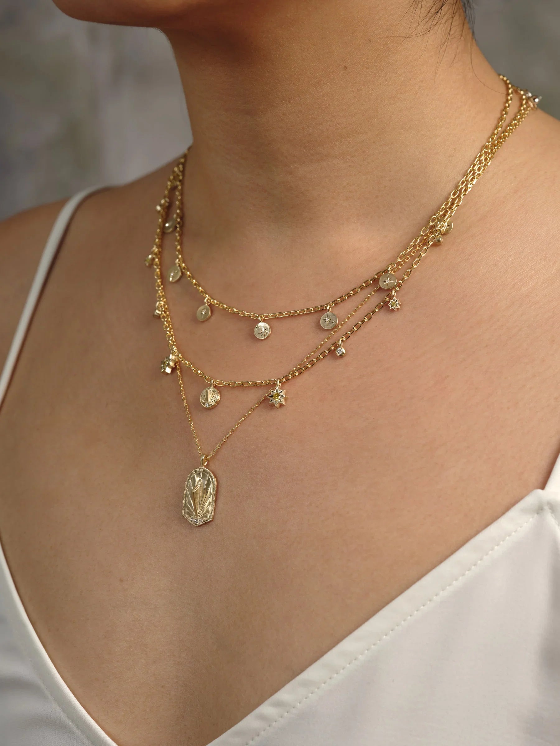 Close-up of layered Fairmined gold necklaces with hand-engraved celestial charms on natural skin and soft warm light