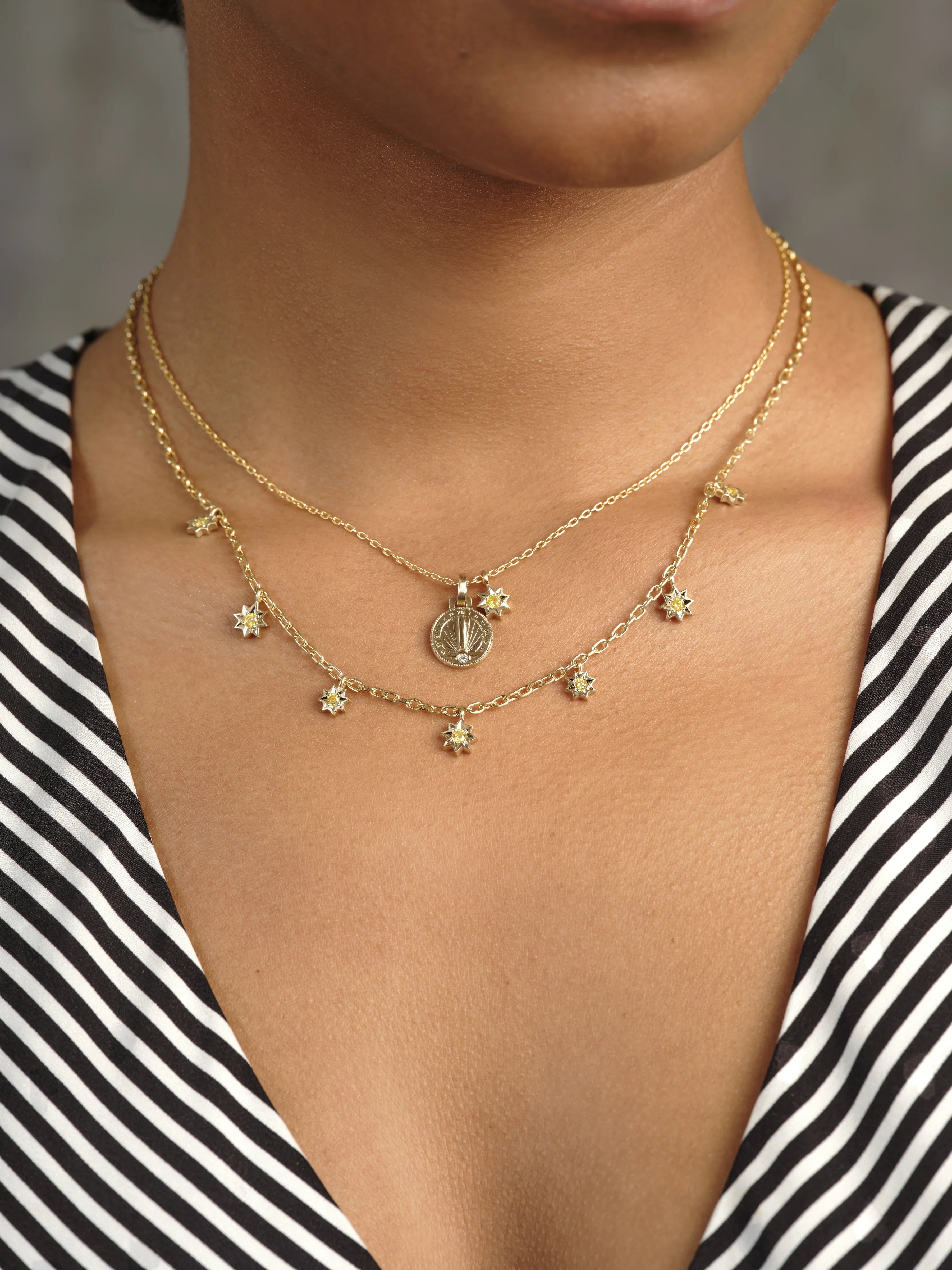 Close-up of layered Fairmined gold sun pendant necklace and star charm chain on warm skin with striped black and white top