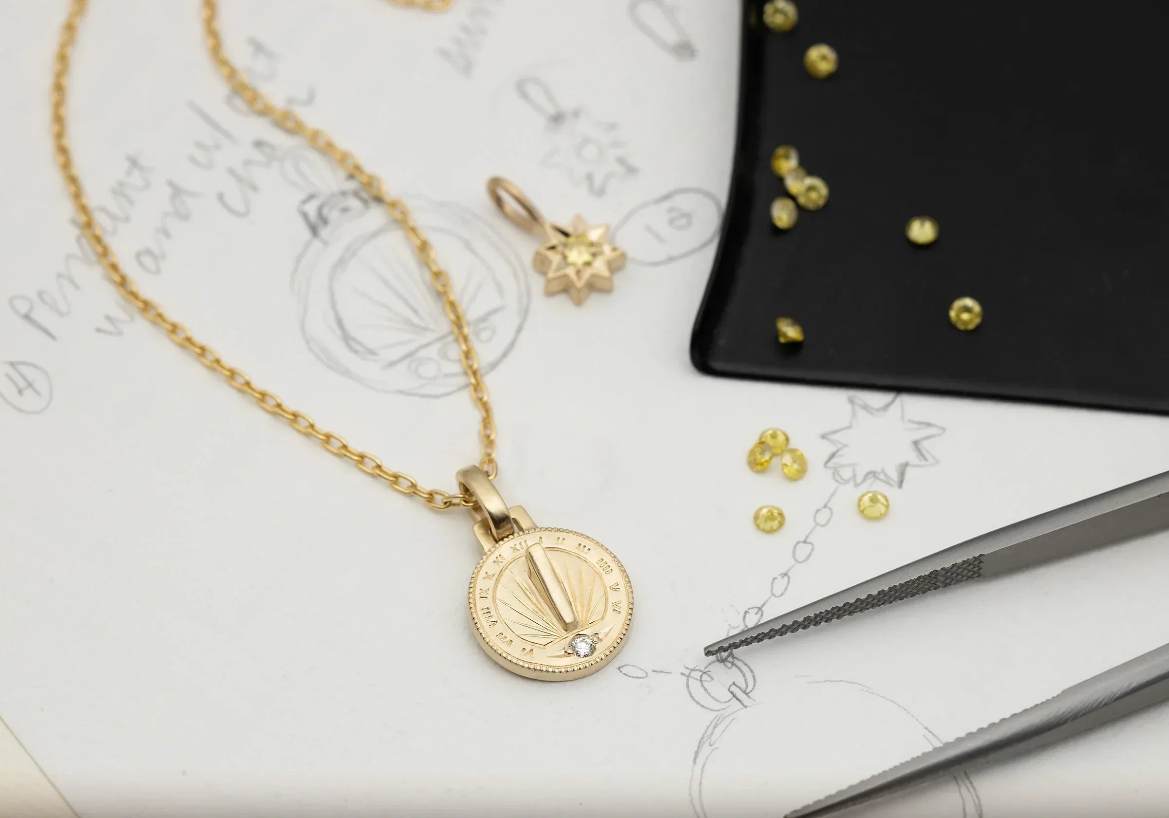 Handcrafted gold sundial pendant necklace with diamond detail on sketch paper with jeweler's tweezers and yellow gems