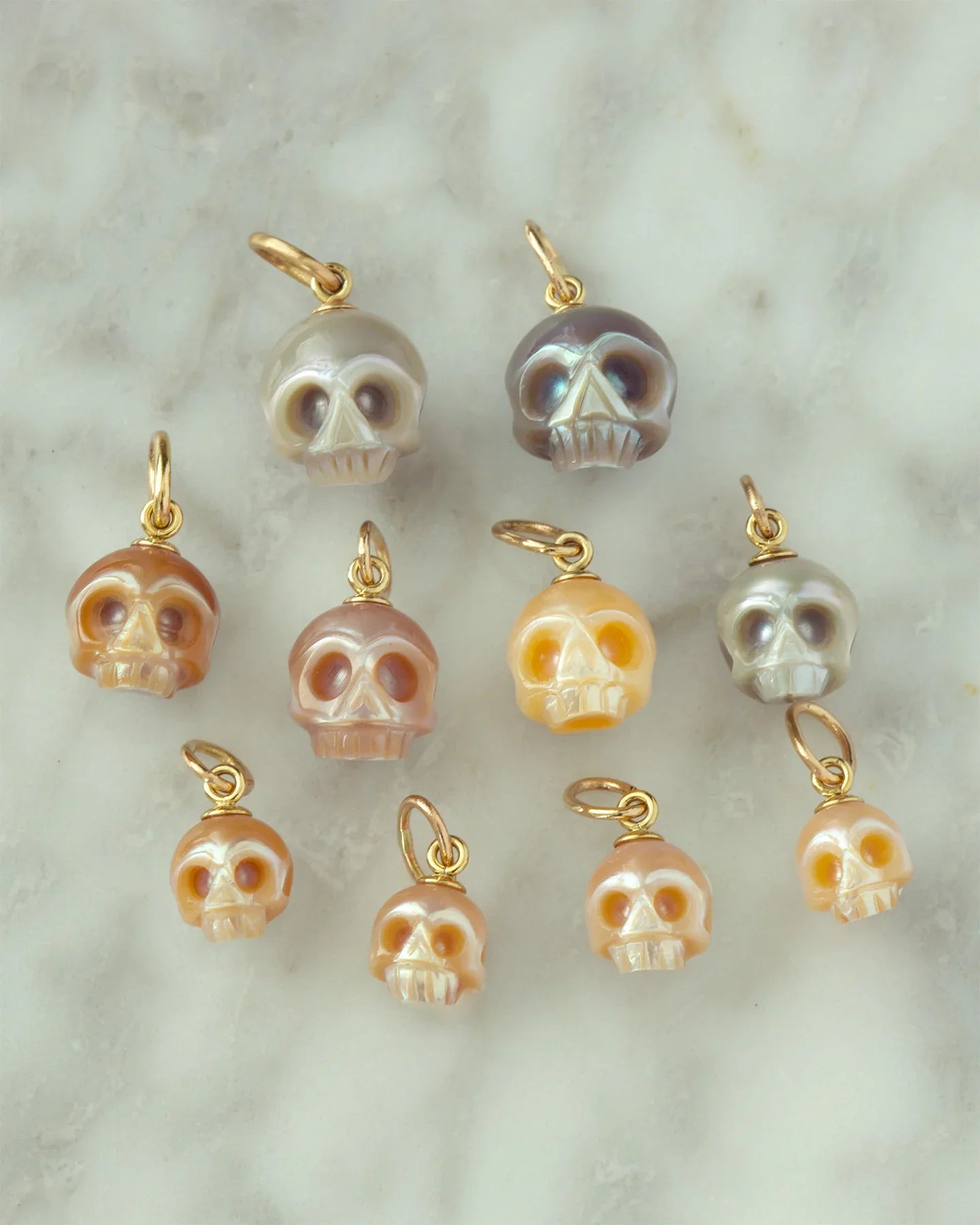Close-up of ten hand-fabricated pearl skull charms with gold loops on smooth marble surface