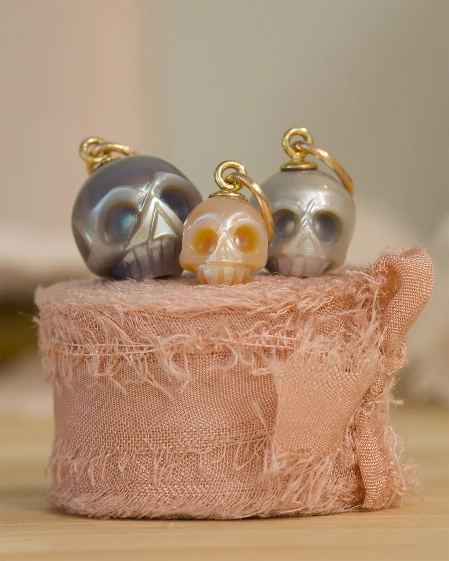 Three hand-carved pearl skull pendants with gold loops, displayed on textured blush fabric, warm natural light
