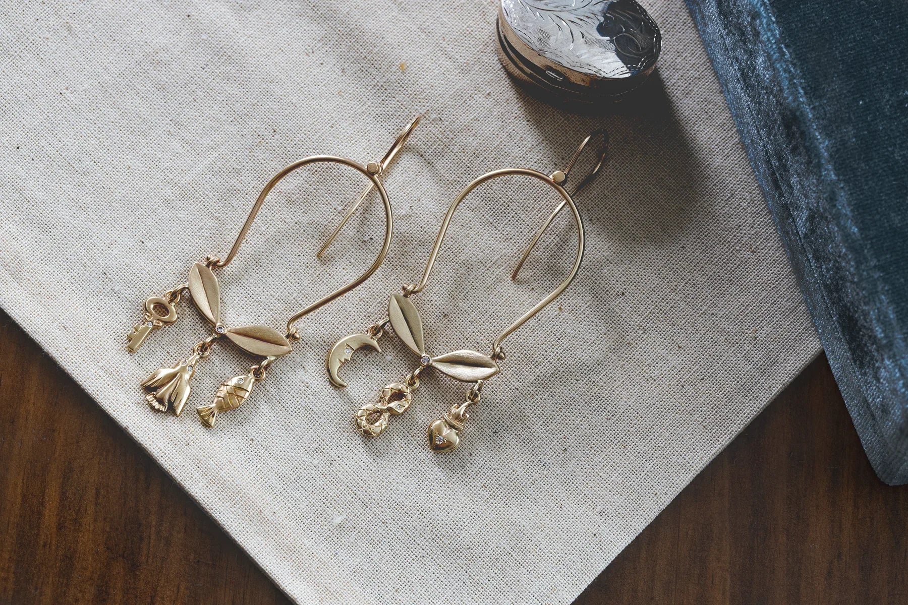 Handcrafted gold earrings with charms including key, flower, fish, moon, infinity, and heart on natural linen
