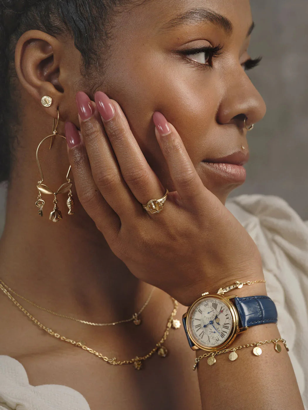Woman wearing handcrafted gold jewelry including earrings, moon phase ring, layered necklaces, and vintage watch with blue leather strap