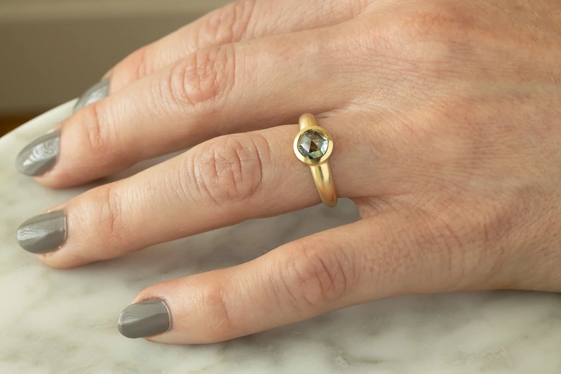 Hand wearing Fairmined 14k gold ring with bezel-set green sapphire, natural light on marble surface