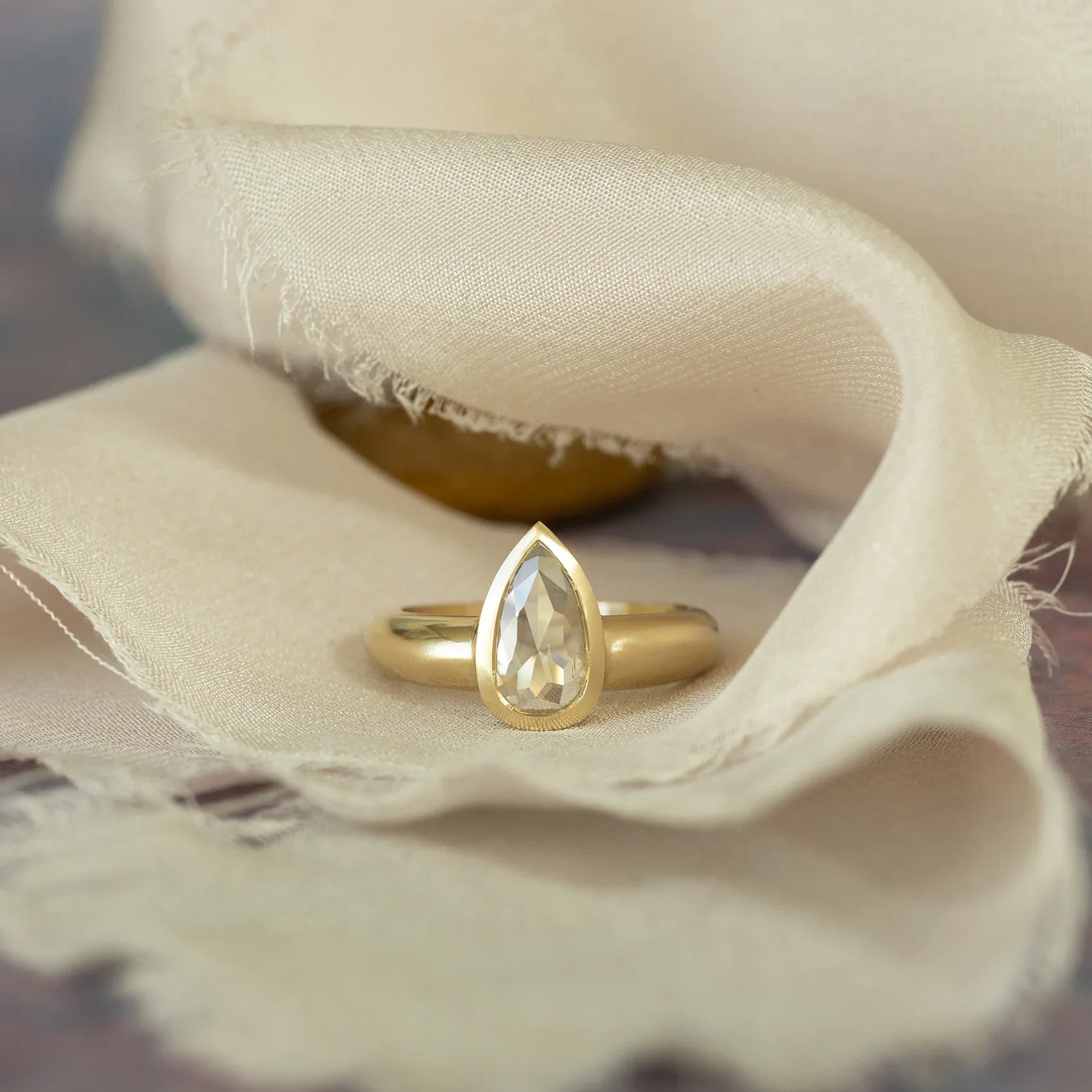 Close-up of gold ring with pear-shaped clear gemstone, soft natural light on beige fabric