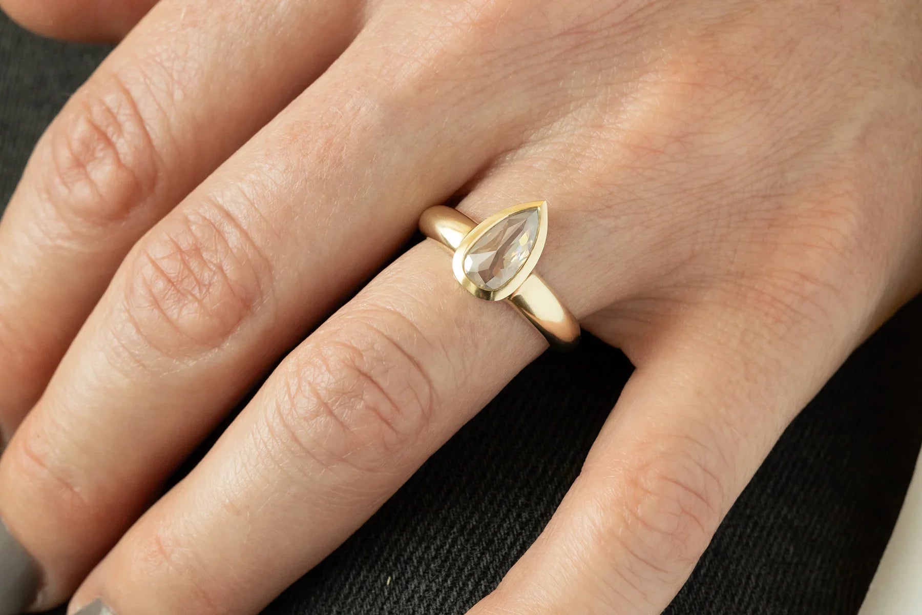 Close-up of hand wearing 14k yellow gold ring with pear-shaped faceted diamond, soft natural light