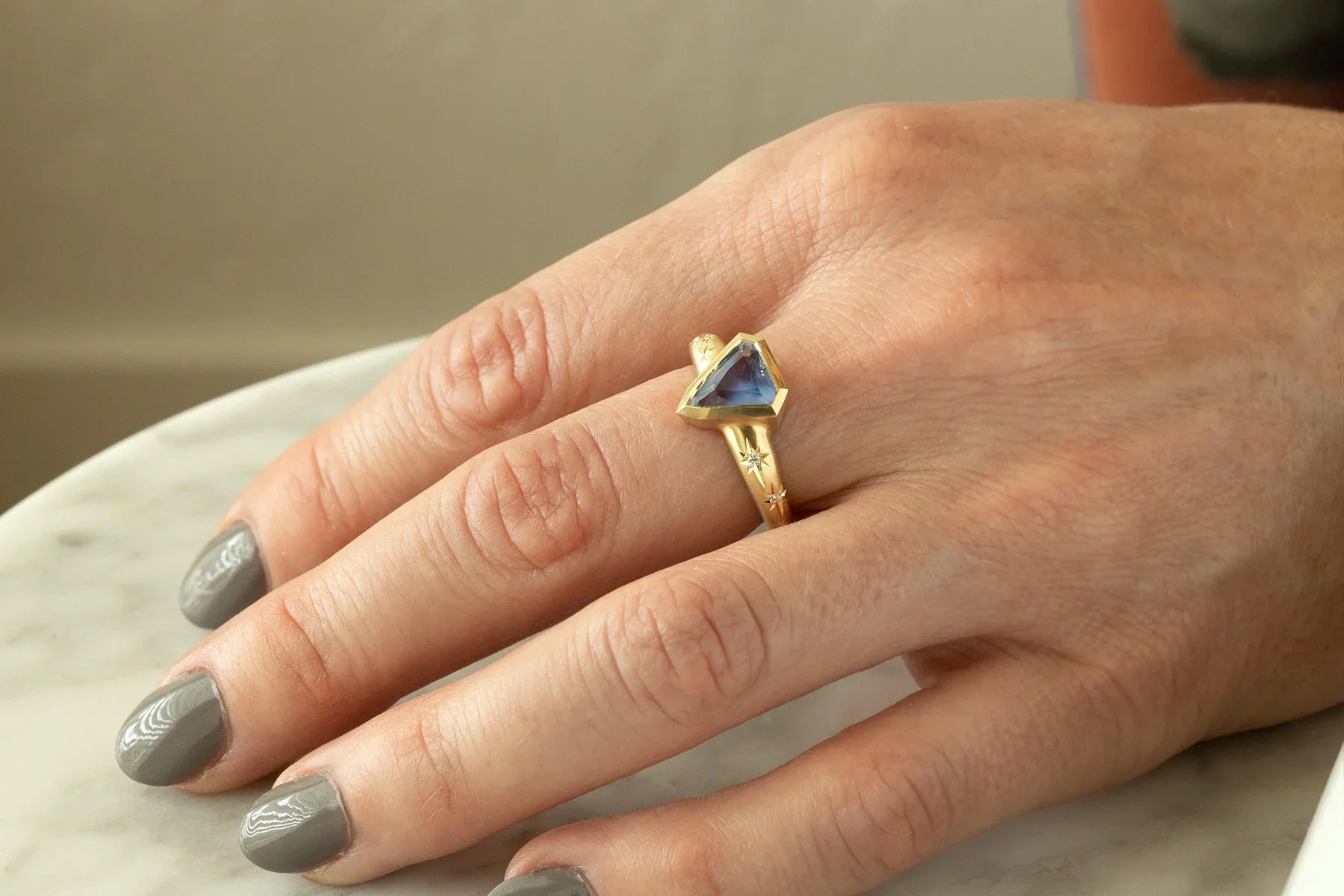 Hand wearing 14k yellow gold ring with triangular blue sapphire and hand-engraved star details on matte marble surface
