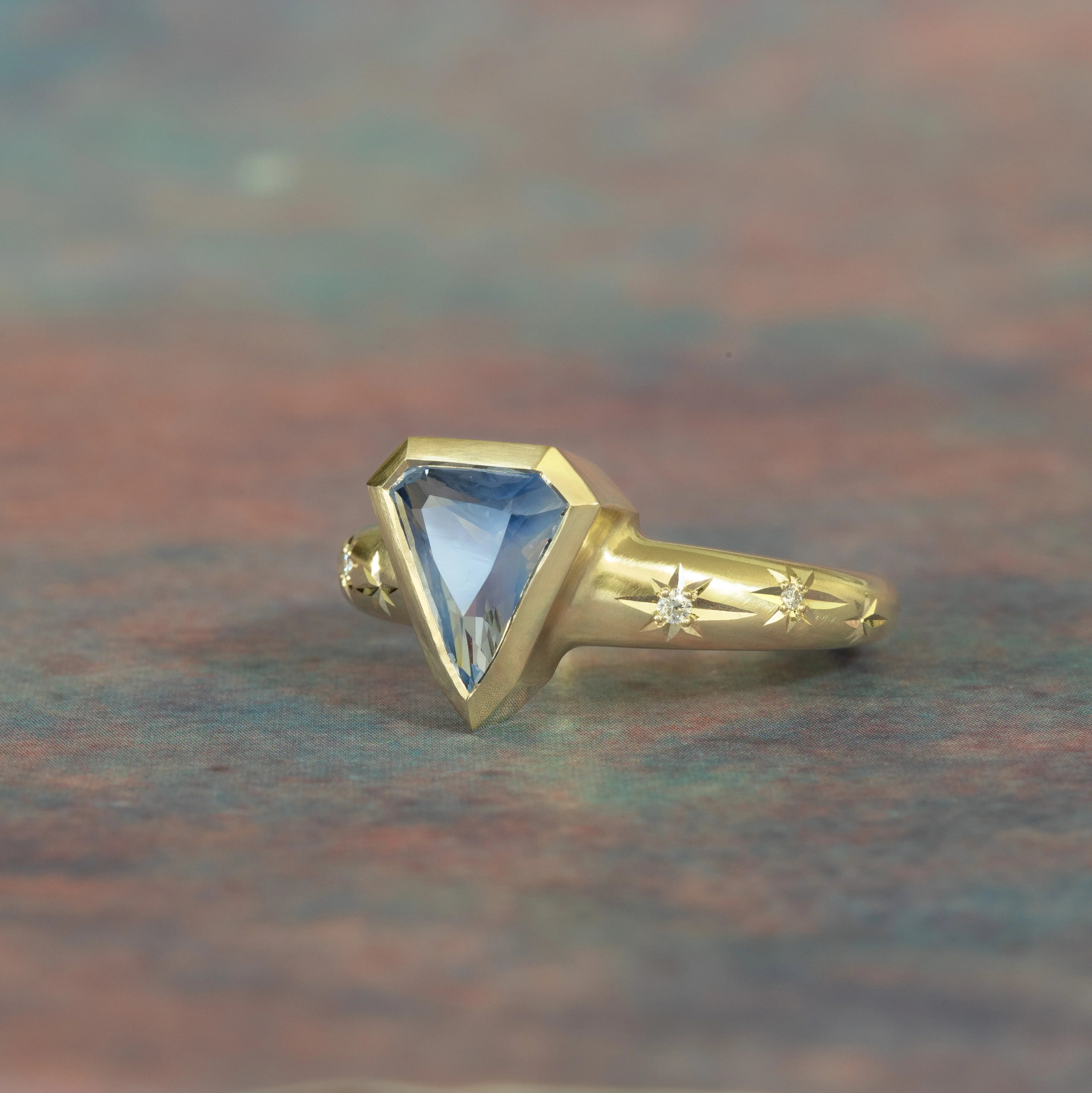 Hand-fabricated 14k gold ring with shield-shaped sapphire and star-engraved diamond accents on soft textured background