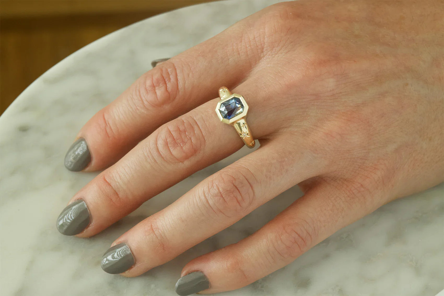 Hand wearing 14k gold ring with faceted blue sapphire and delicate star engravings on band, on marble surface
