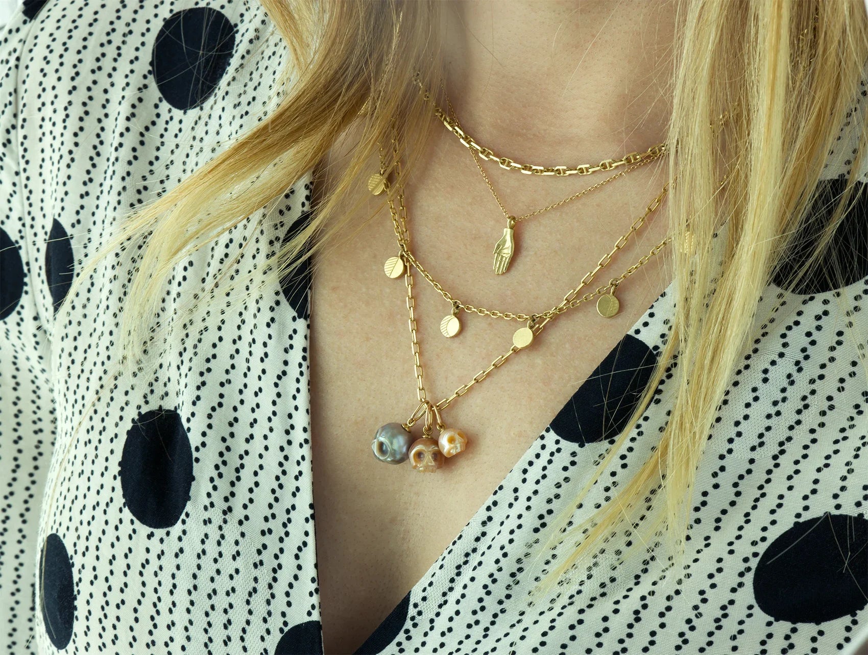 Close-up of layered handcrafted gold necklaces with three pearl skull pendants worn on a woman with blonde hair in soft natural light