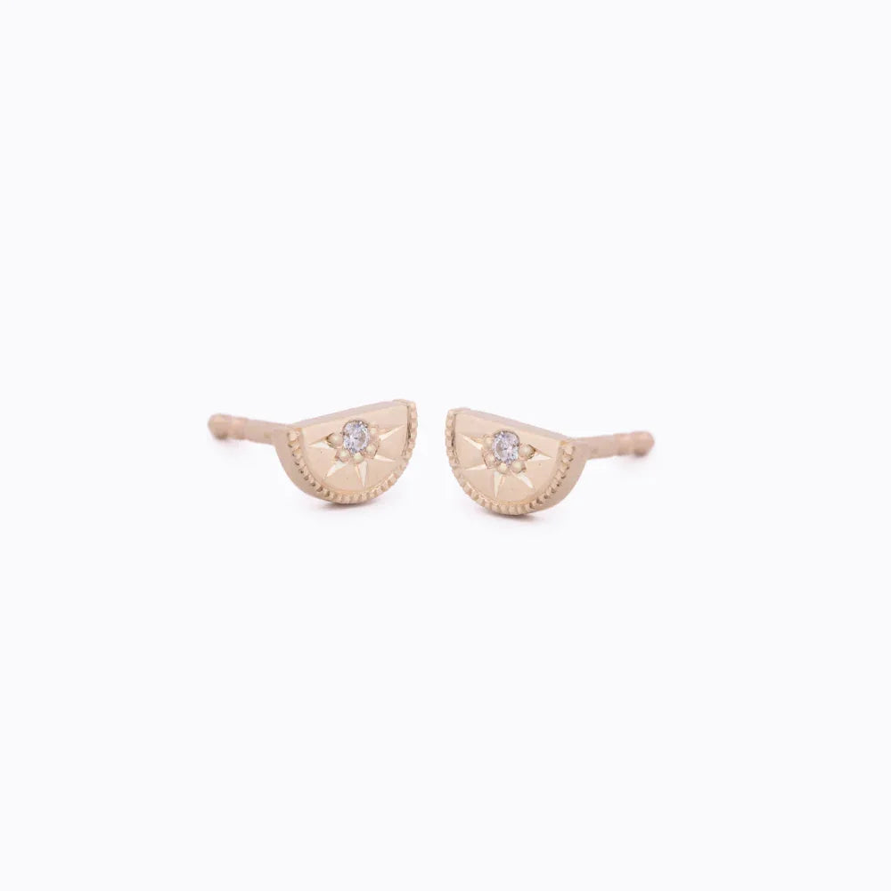 Close-up of delicate 14k gold half-moon stud earrings with central diamond and hand-engraved sunburst detail on white background