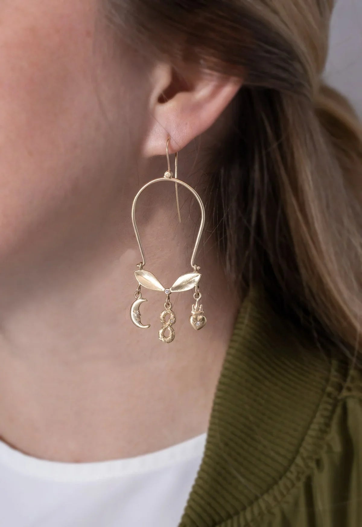 Close-up of hand-fabricated gold earring with moon, infinity, and heart charms worn against soft natural skin tones