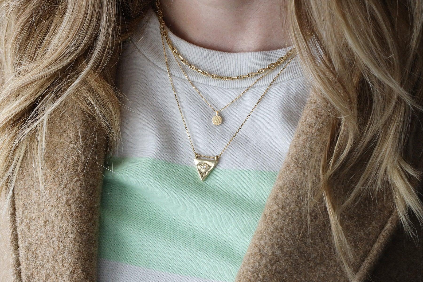 Layered gold necklaces including a triangle pendant with an eye motif, worn with a beige coat and striped sweater