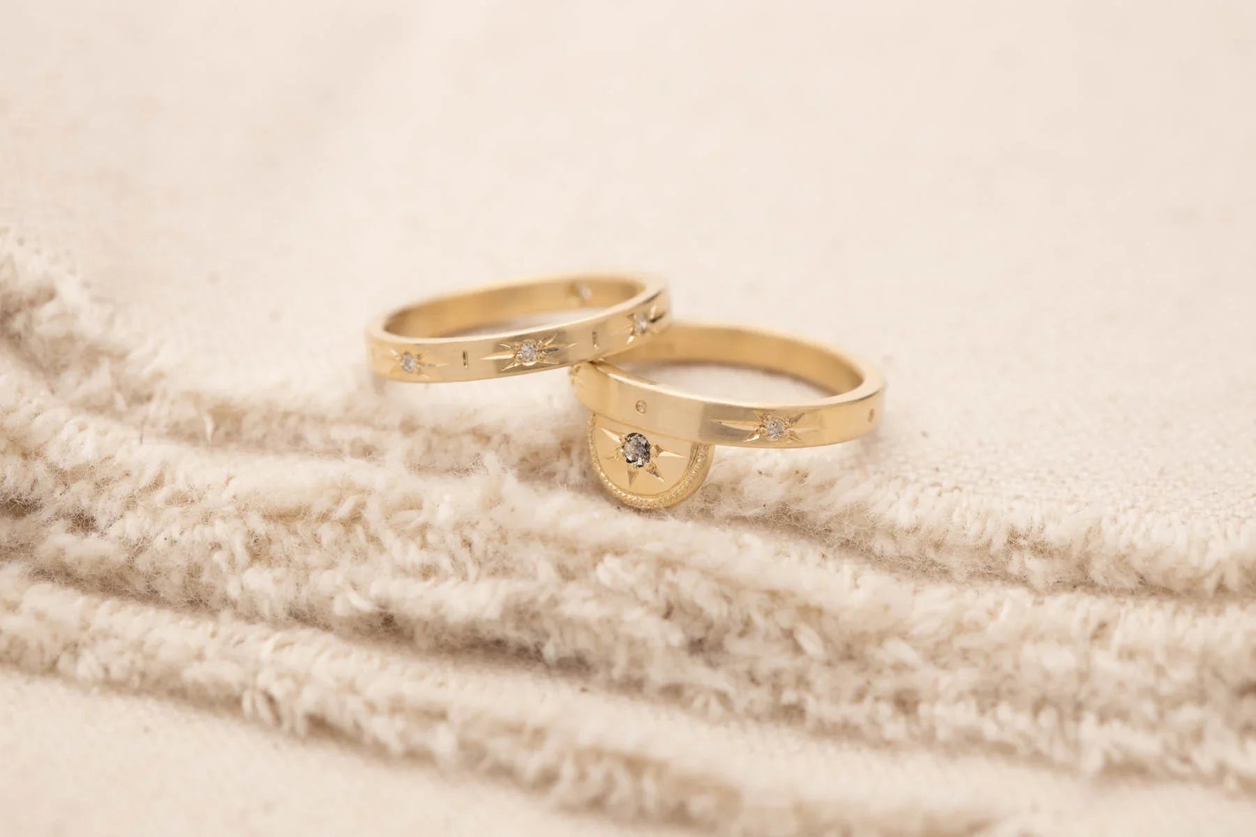 Two handcrafted gold rings with starburst diamond accents on soft beige textured fabric, warm natural light